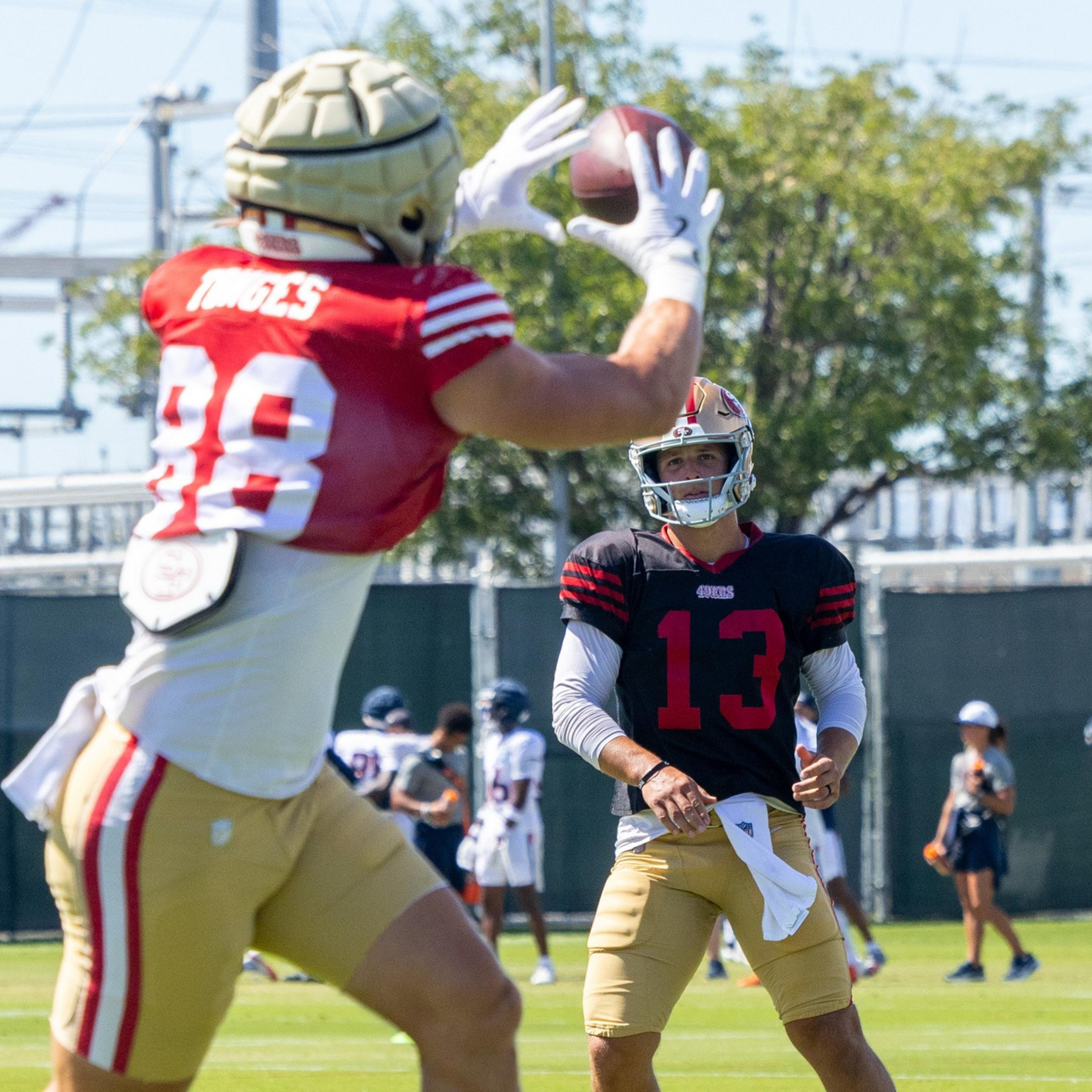 A football player in a red jersey and gold helmet catches a ball while another player in a black jersey and gold helmet watches during practice.