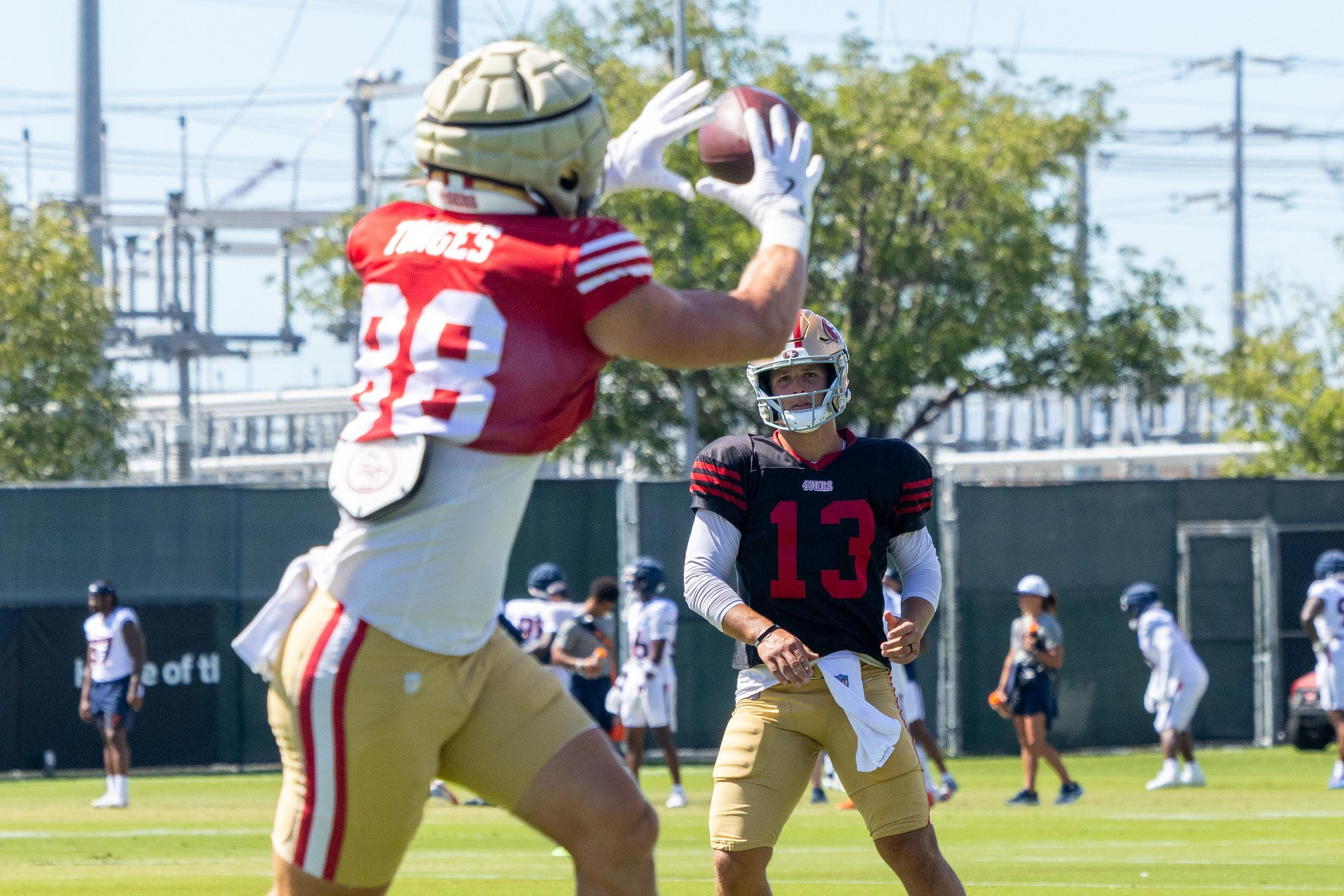 A football player in a red jersey and gold helmet catches a ball while another player in a black jersey and gold helmet watches during practice.