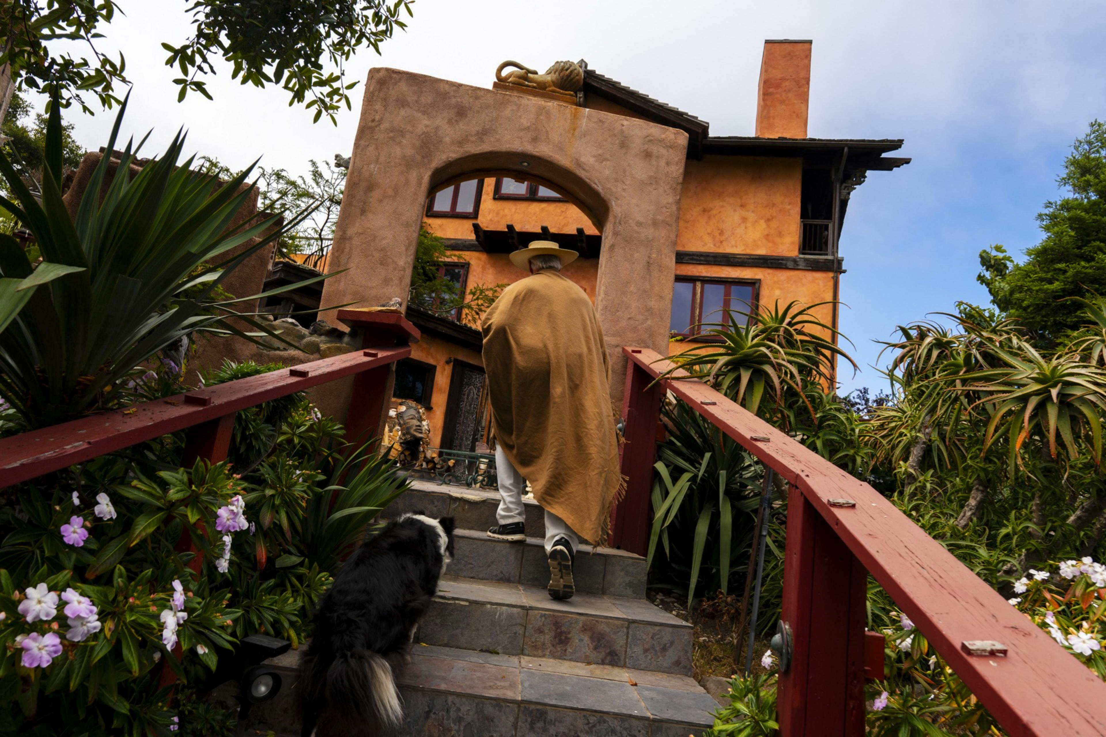 A person in a brown cloak and hat climbs stone steps next to a dog. They approach an orange house with lush greenery and a wooden archway.