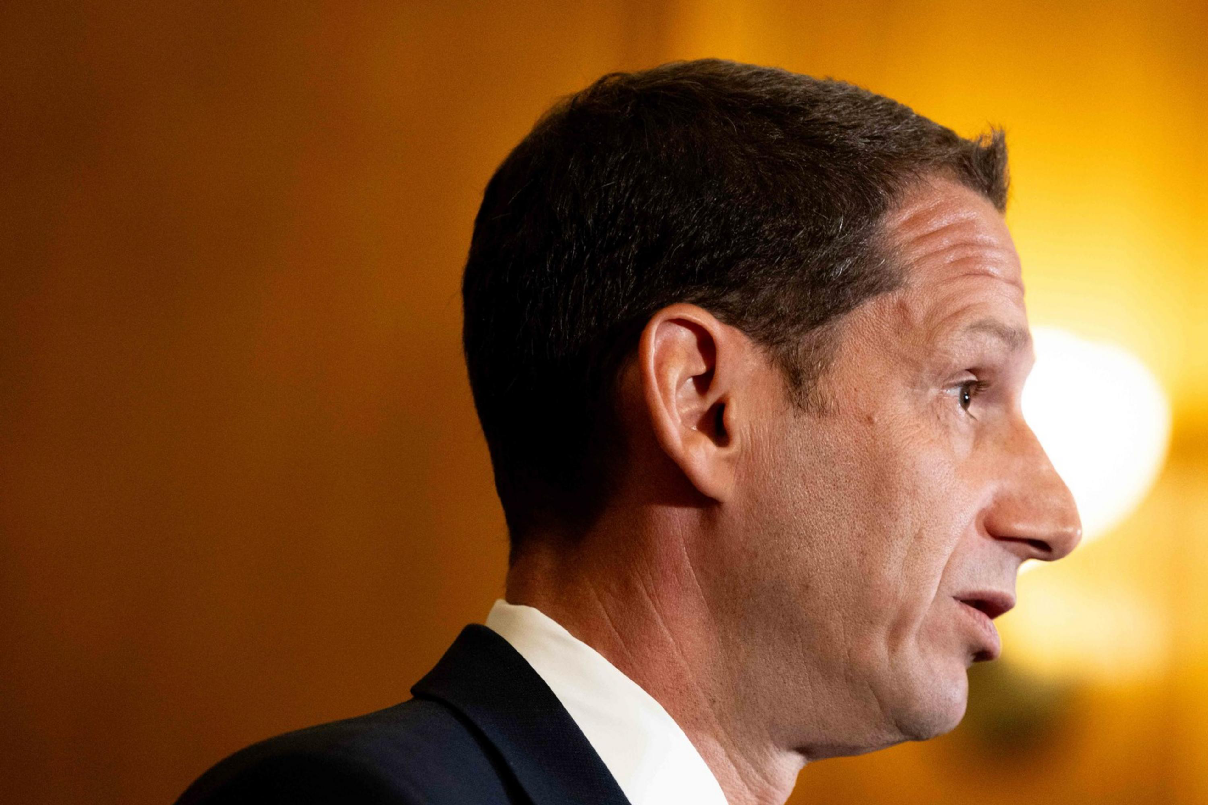 The image shows a person in profile speaking into a microphone. They are wearing a suit and tie. The background is warmly lit and slightly blurred.