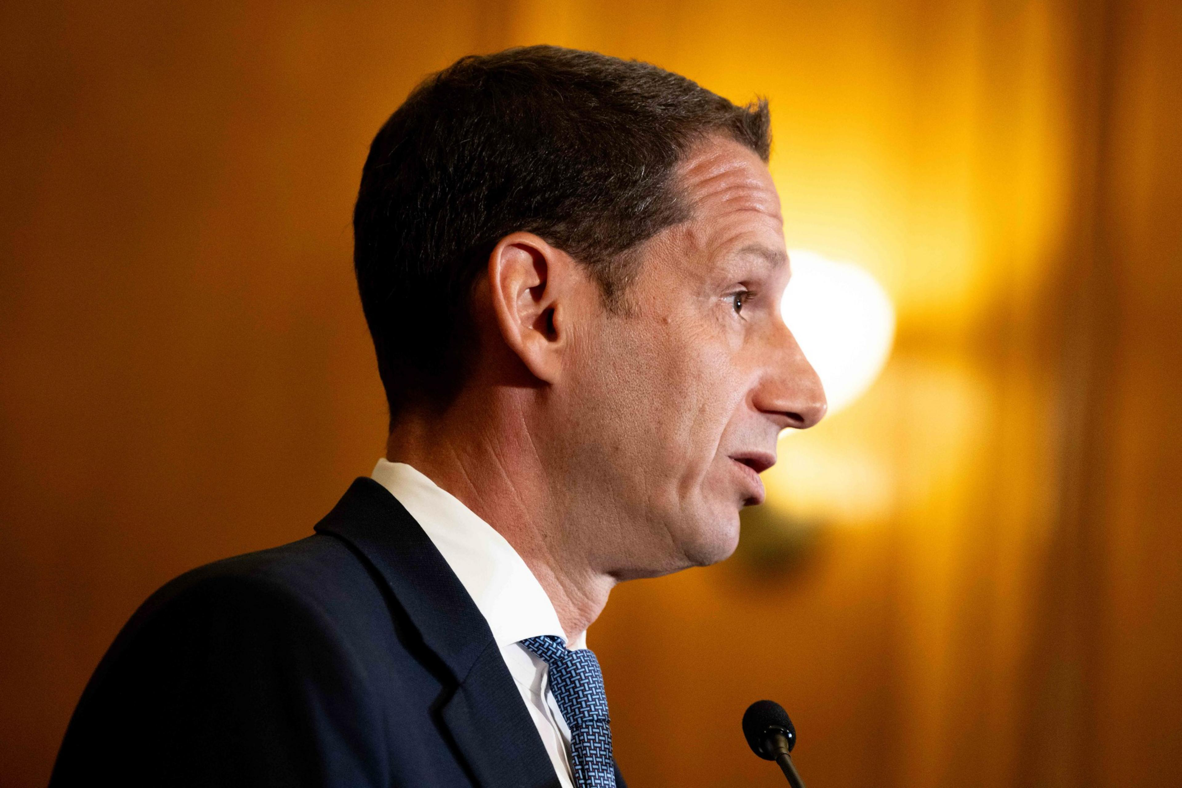 The image shows a person in profile speaking into a microphone. They are wearing a suit and tie. The background is warmly lit and slightly blurred.