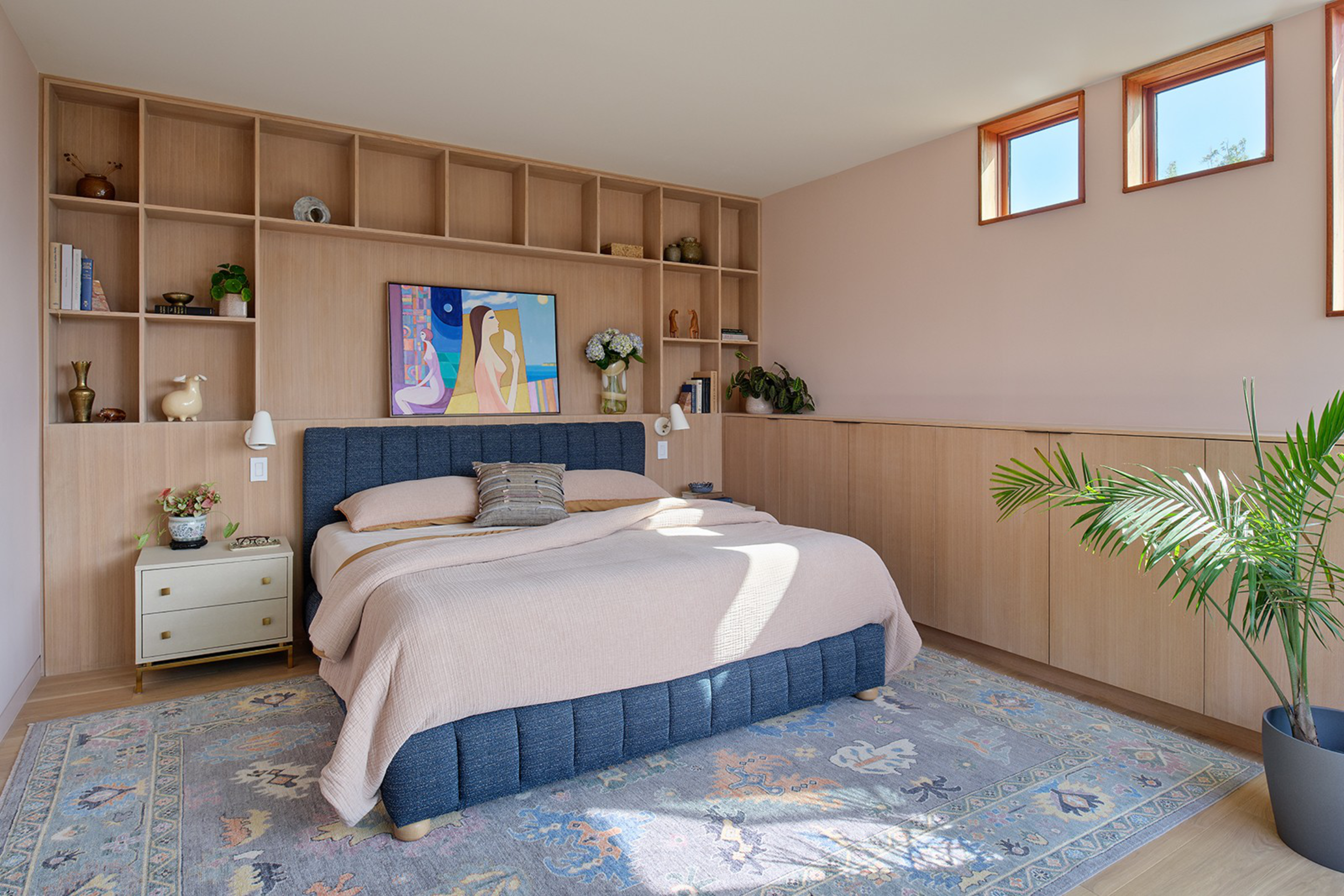 A modern bedroom with a blue upholstered bed, beige bedding, wooden shelves filled with decor, a floral rug, and three small high windows letting in light.