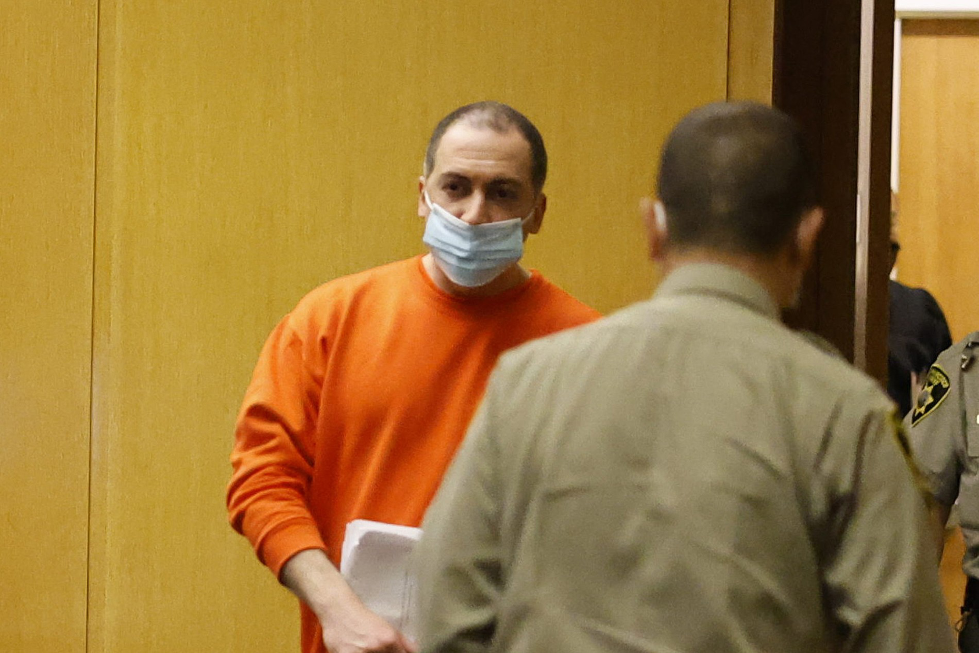 A man in an orange shirt and a blue face mask holds papers while a uniformed officer faces away from the camera in a wood-paneled room.