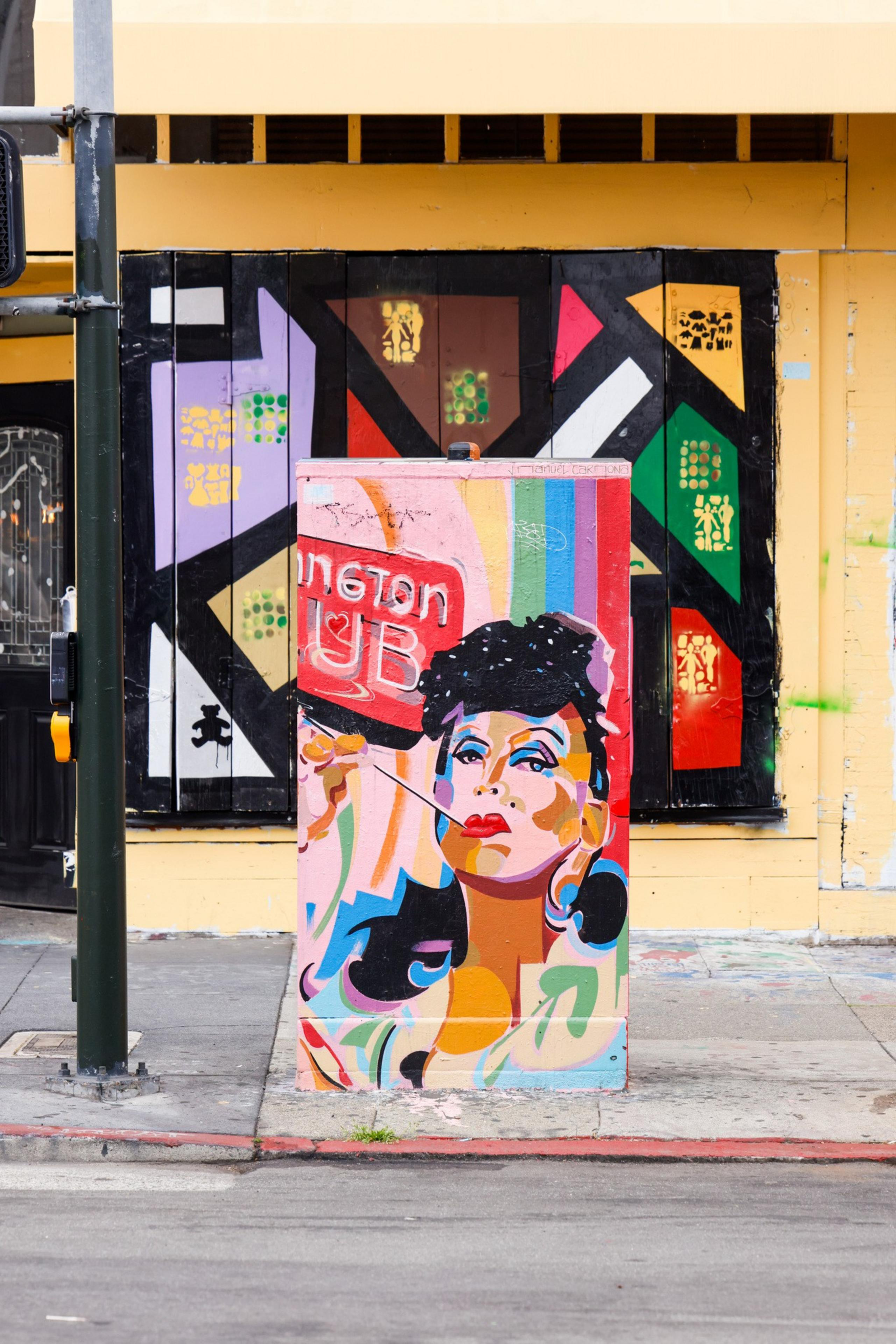 A colorful street mural features a stylized portrait of a person with bold red lips, black hair, and vivid abstract shapes. Vibrant colors create a lively backdrop.