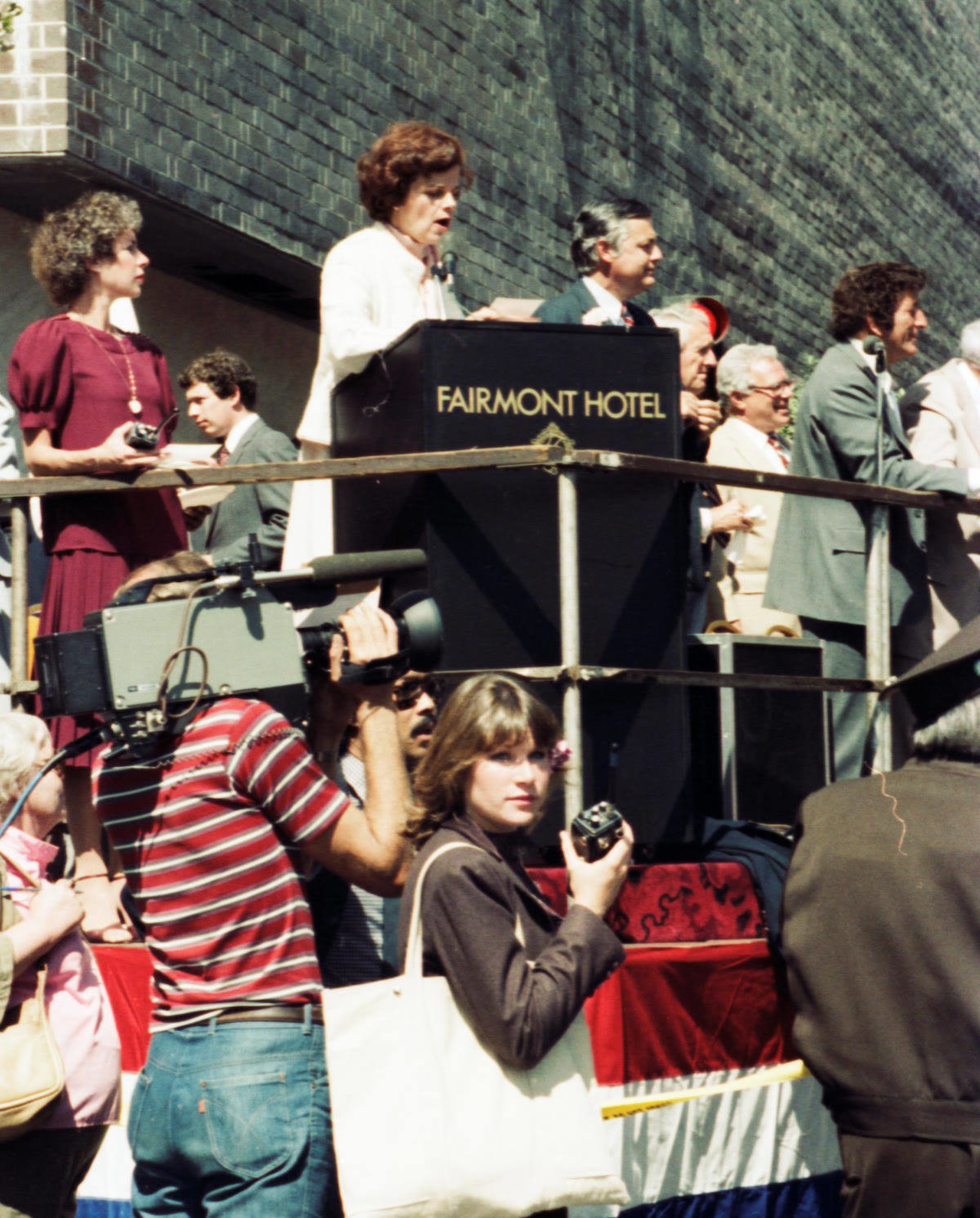A woman speaks at a podium labeled "Fairmont Hotel" surrounded by people, while a cameraman and a woman with a camera stand in the foreground.