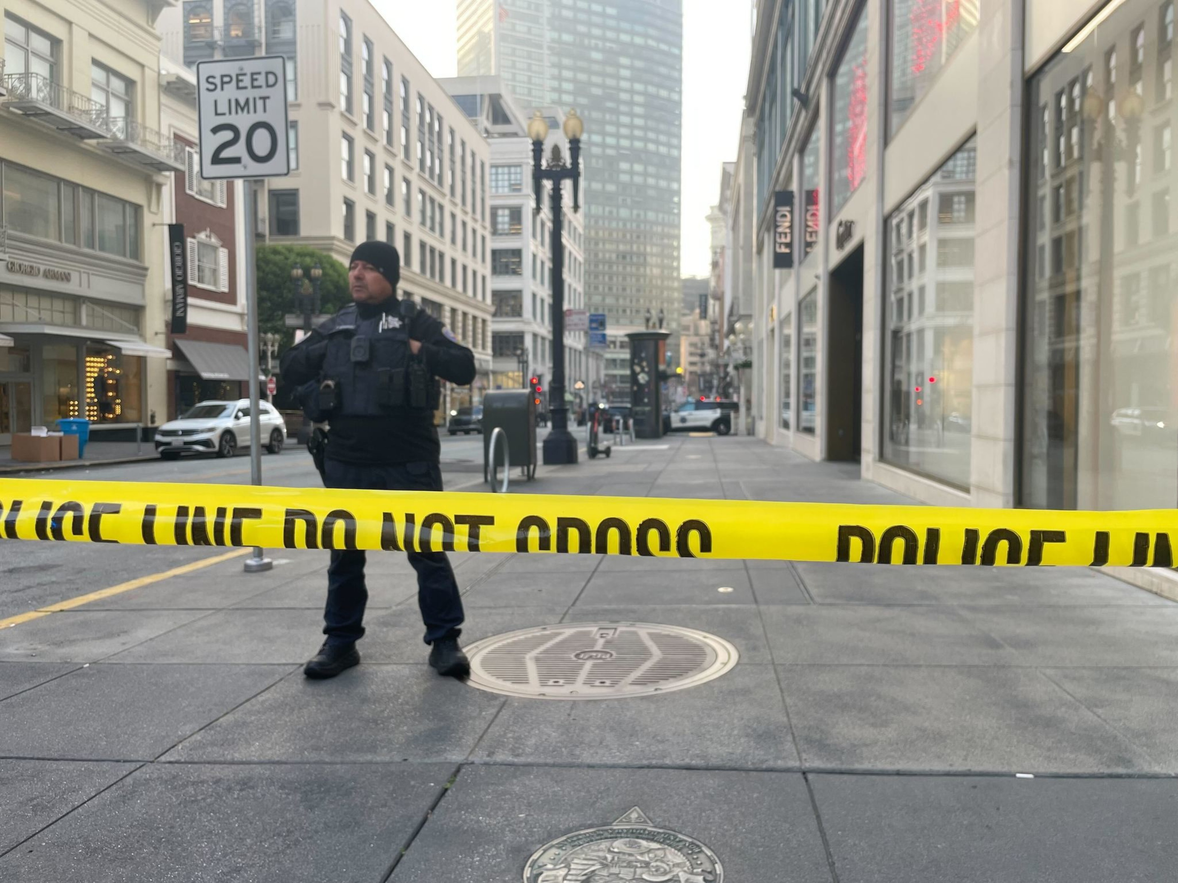 A police officer stands behind yellow &quot;Police Line Do Not Cross&quot; tape on a city street. Nearby is a speed limit sign, and tall buildings line the background.