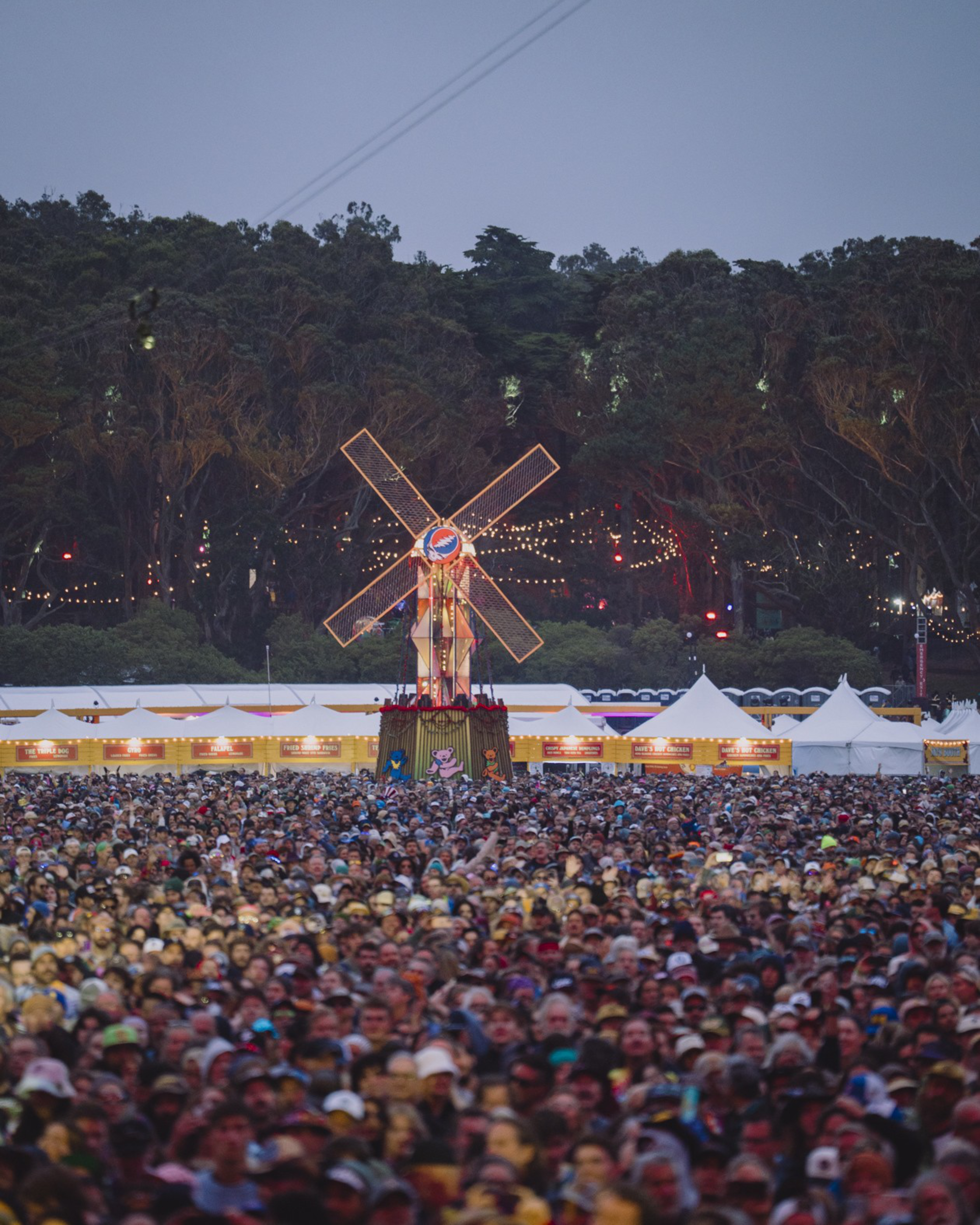 A large crowd gathers outdoors at dusk in front of a lit windmill structure, with tents and trees decorated with string lights in the background.