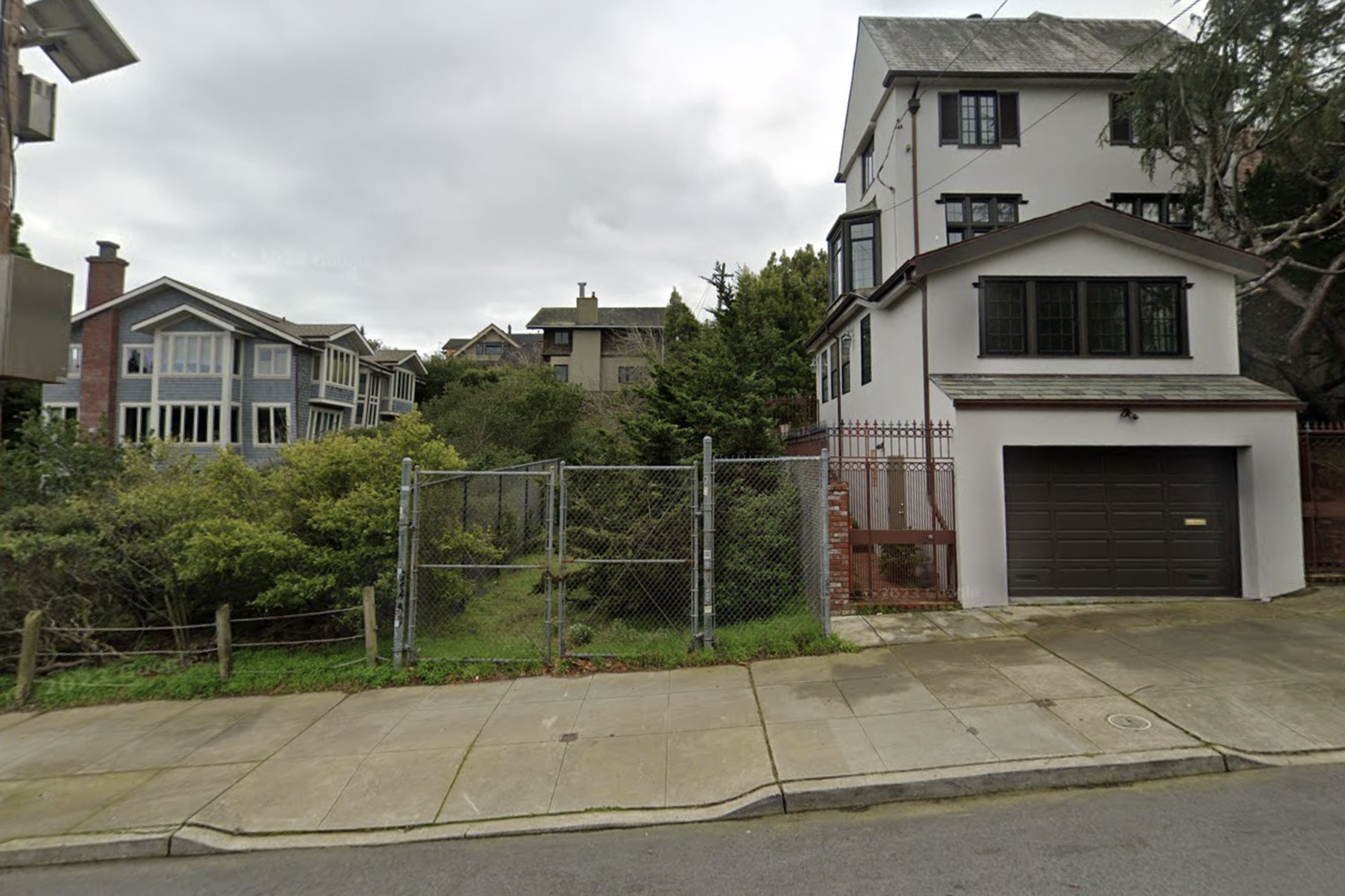 A narrow, gated, overgrown lot sits between a white multi-story house with a dark garage door and a row of houses with large windows on a cloudy day.
