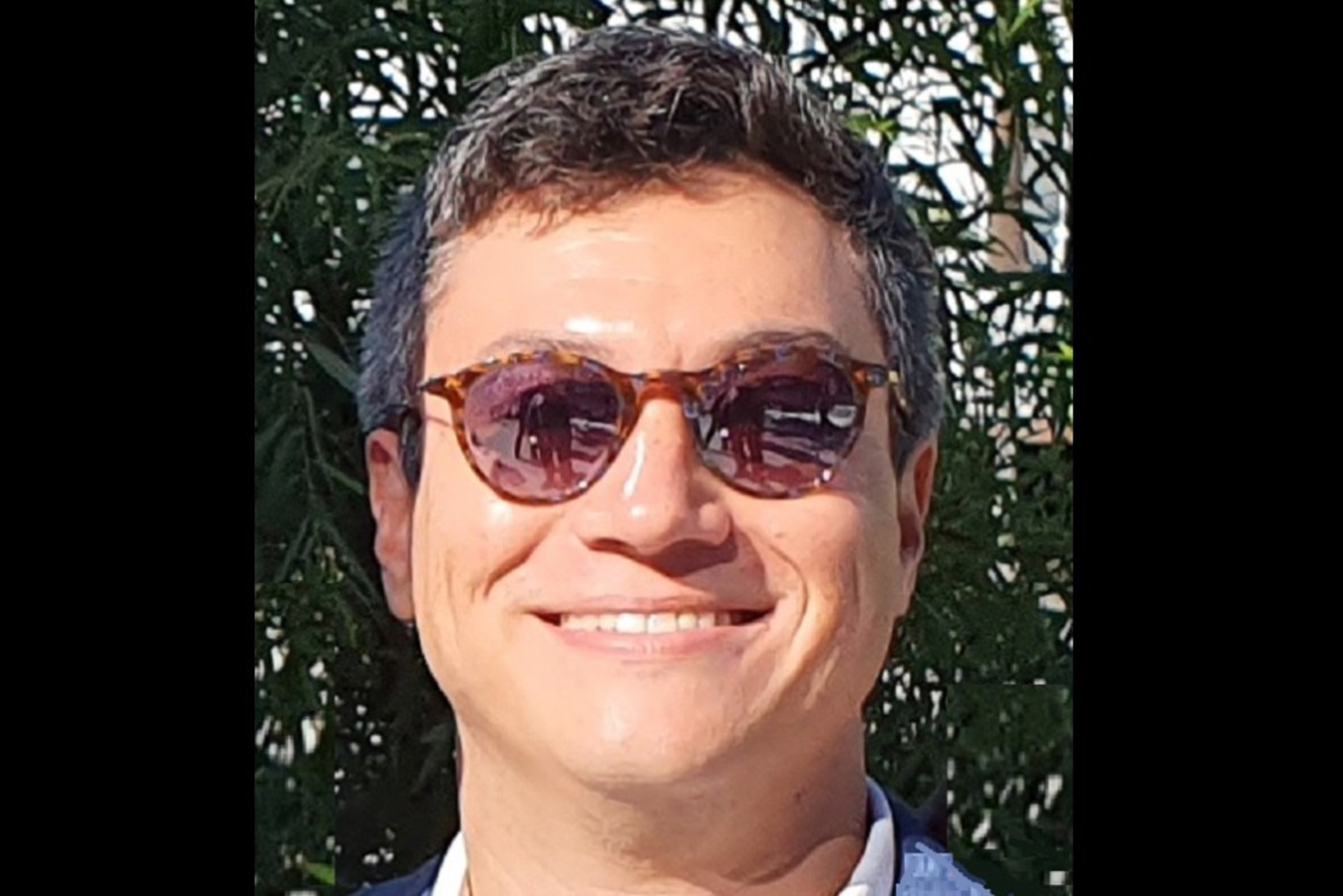 A man with short dark hair is smiling and wearing brown tortoiseshell sunglasses and a blue blazer, standing outdoors with green foliage behind him.