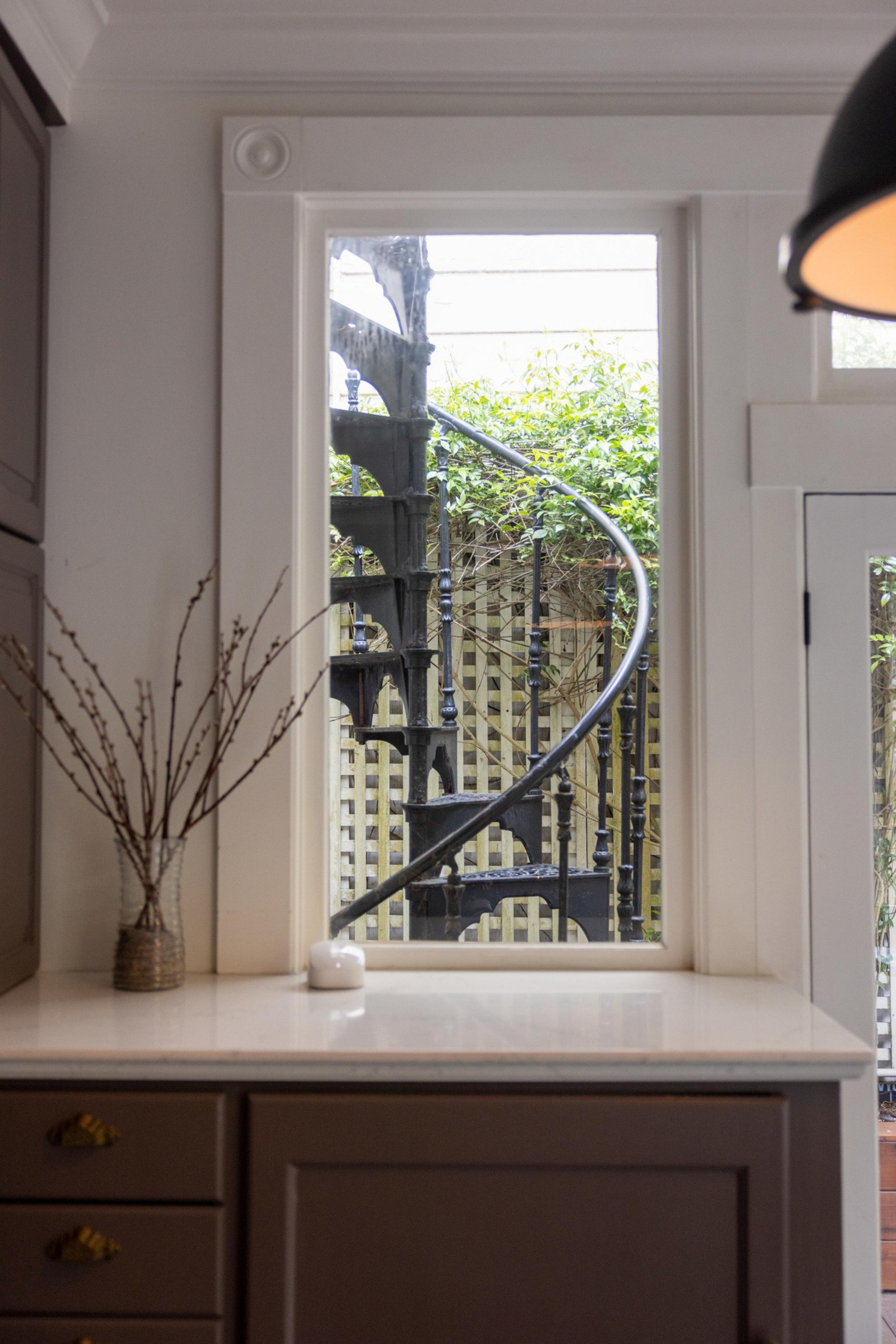 A black spiral staircase is seen through a window in a room with dark cabinets, a vase of twigs, and a small candle on a light countertop.