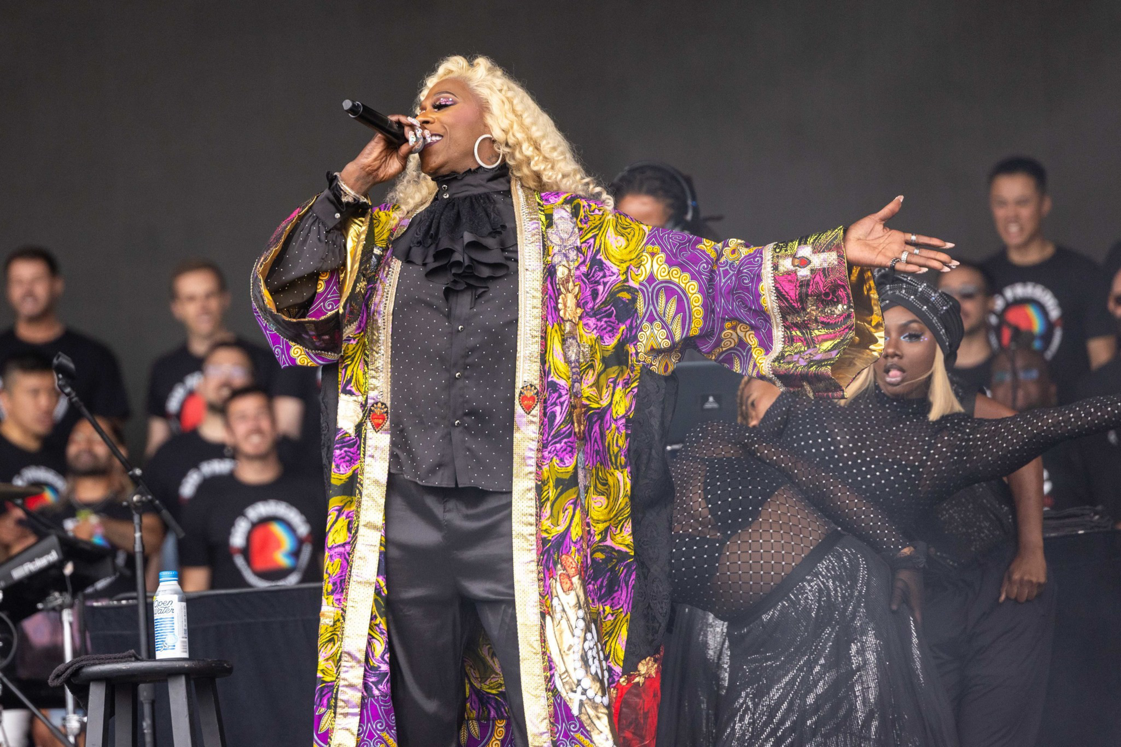 A performer with long blonde hair sings into a microphone wearing a colorful patterned robe over black clothes, while a dancer in black mesh strikes a dramatic pose nearby.