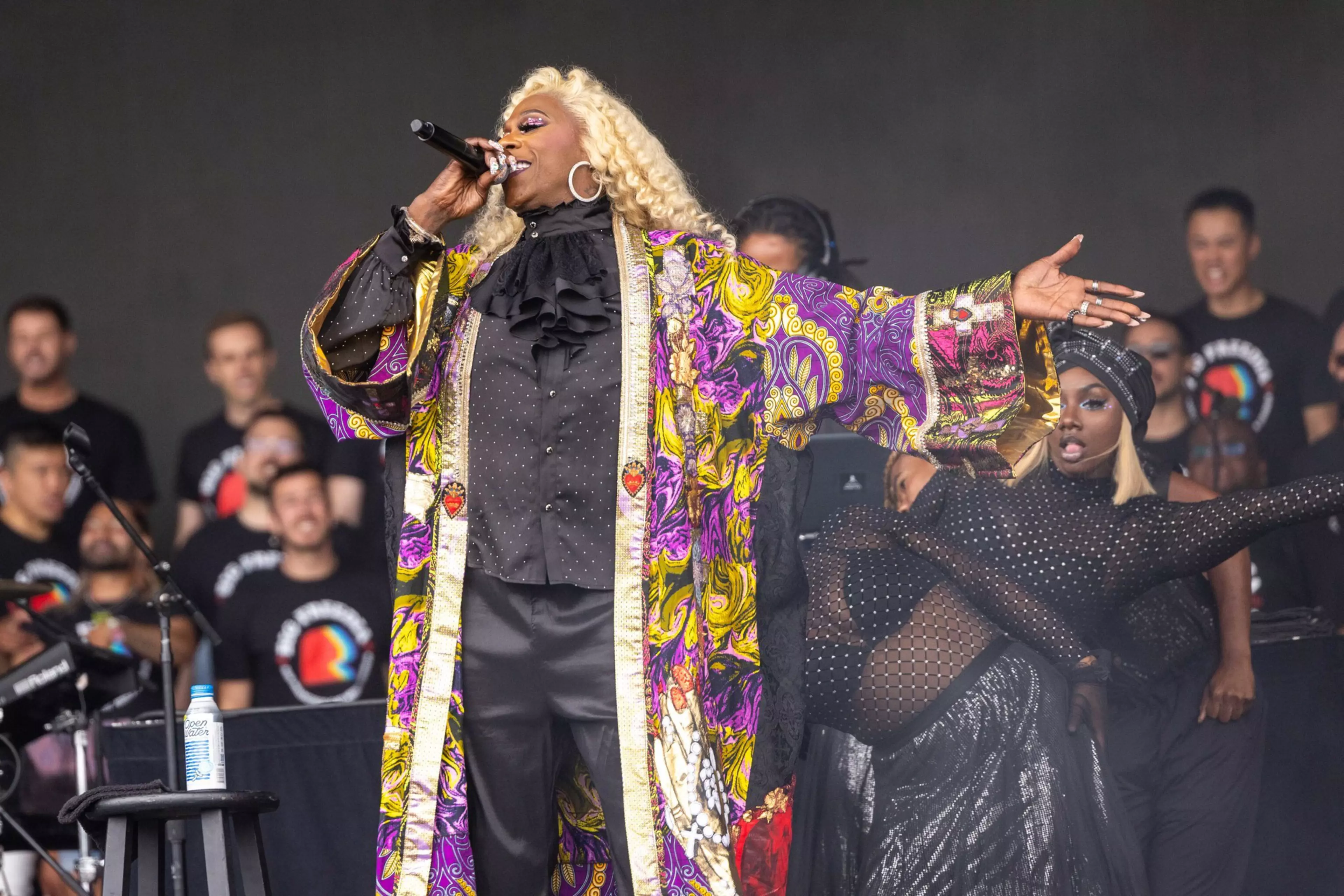 A performer with long blonde hair sings into a microphone wearing a colorful patterned robe over black clothes, while a dancer in black mesh strikes a dramatic pose nearby.
