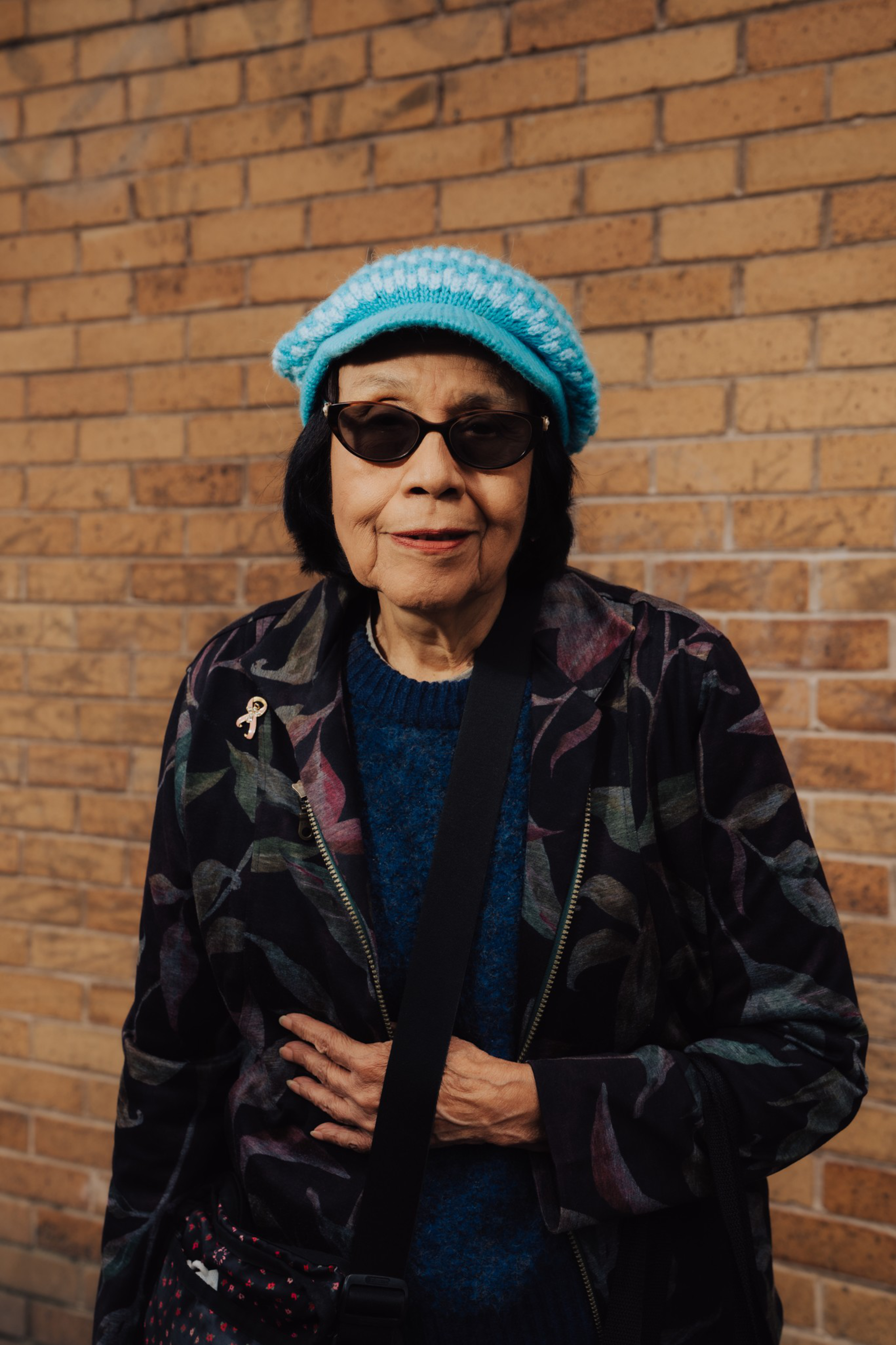 An elderly woman wearing a bright blue knit hat, dark sunglasses, a floral jacket, and a navy sweater stands with her hand on her stomach against a brick wall.