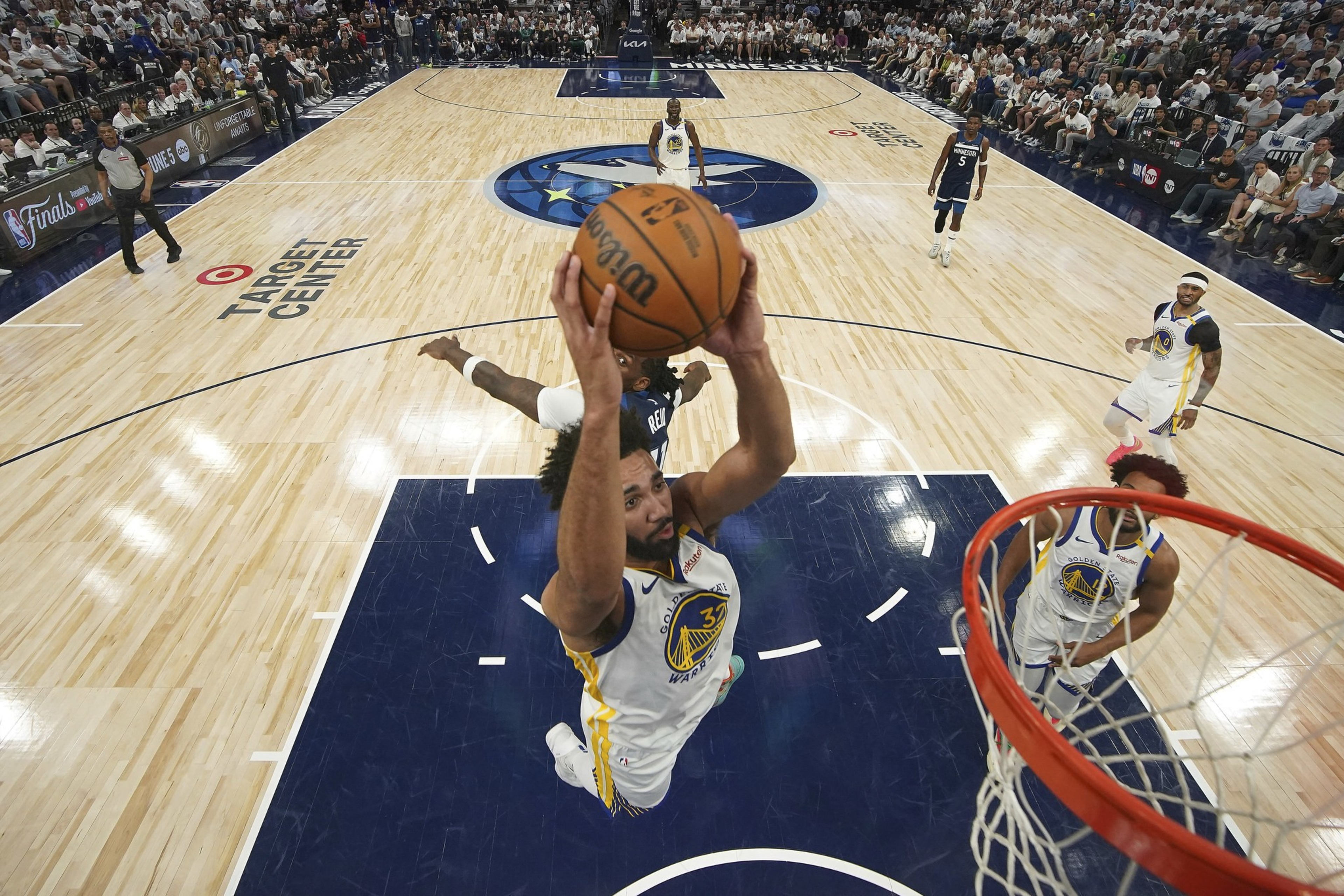 A basketball player in a Warriors jersey jumps to score a basket, while a Timberwolves player attempts to block him on a crowded court at Target Center.
