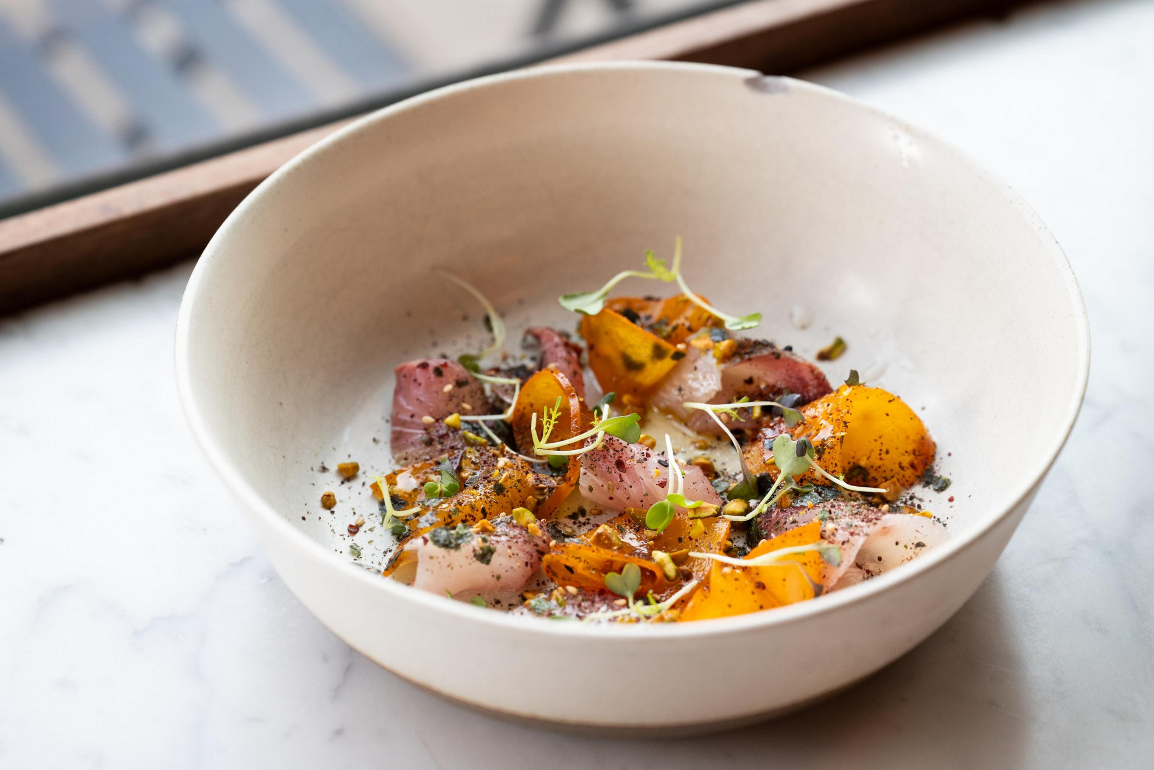 Slices of raw fish are garnished with herbs, spices, and small yellow-orange vegetable pieces in a white bowl on a marble surface.