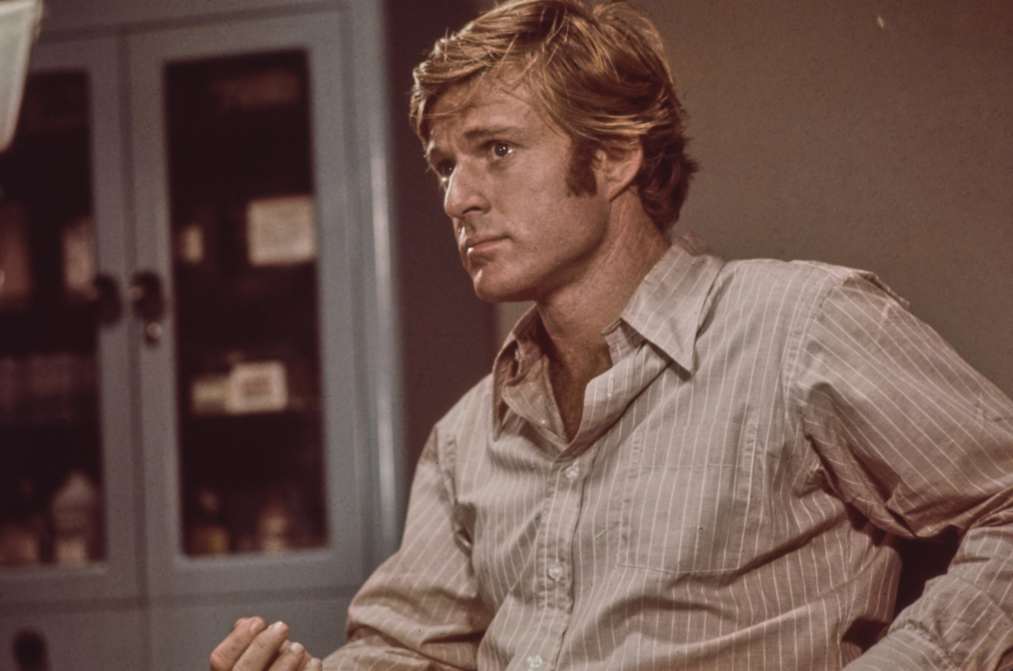 A man with blond hair wears a beige striped shirt, sitting and gesturing with his right hand, looking thoughtfully to the side indoors.
