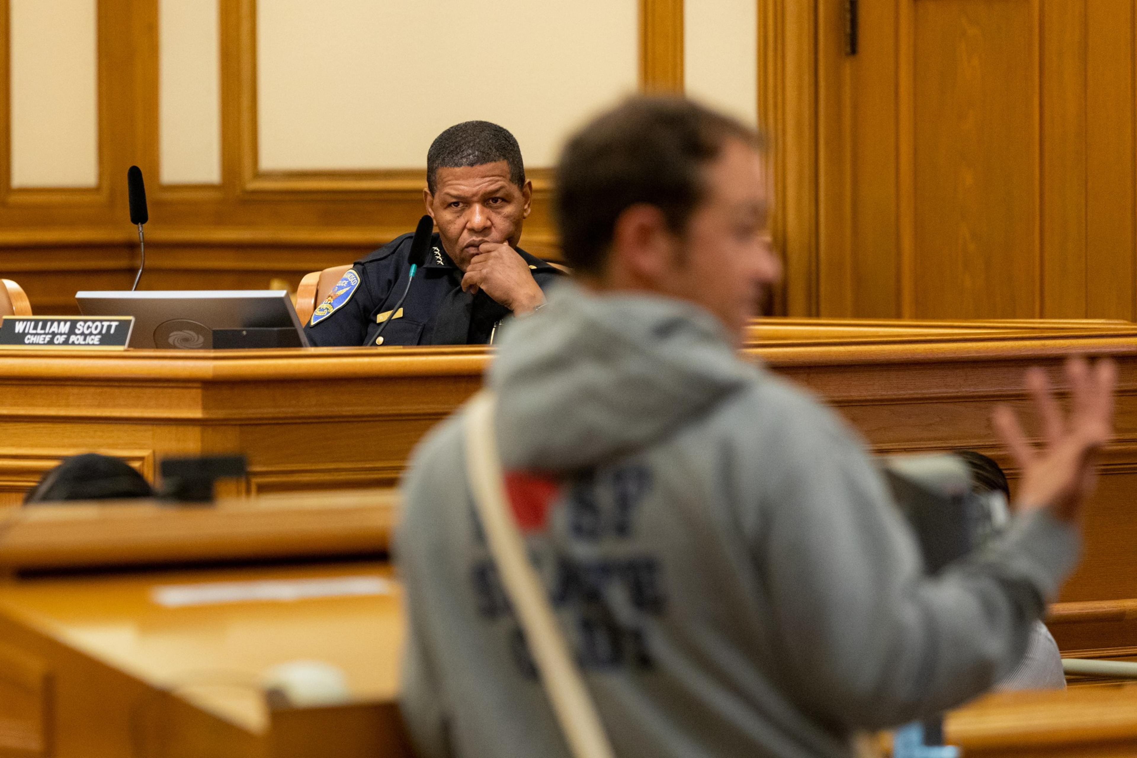 A man in a police uniform labeled "Chief of Police" watches intently as a blurred man gestures while speaking in a wood-paneled room.