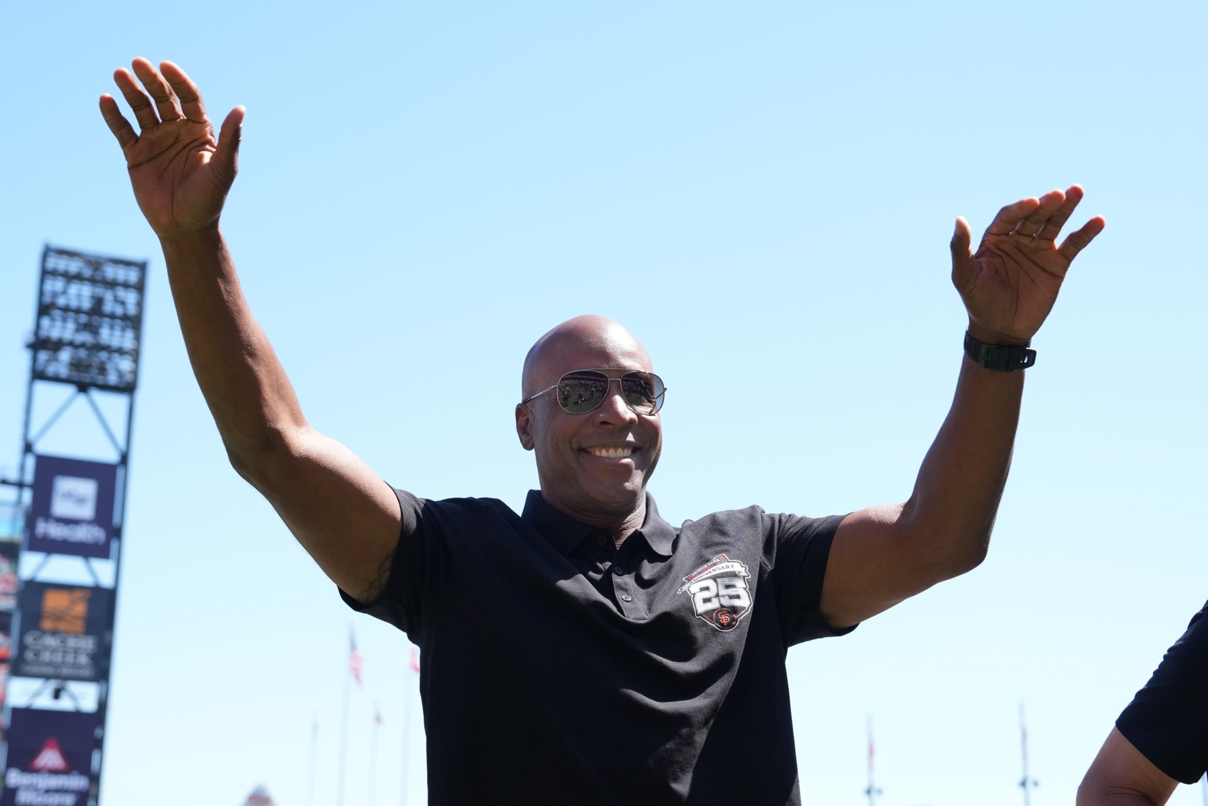 A smiling man in sunglasses and a black shirt raises his arms against a clear blue sky, with stadium signs and flags visible in the background.