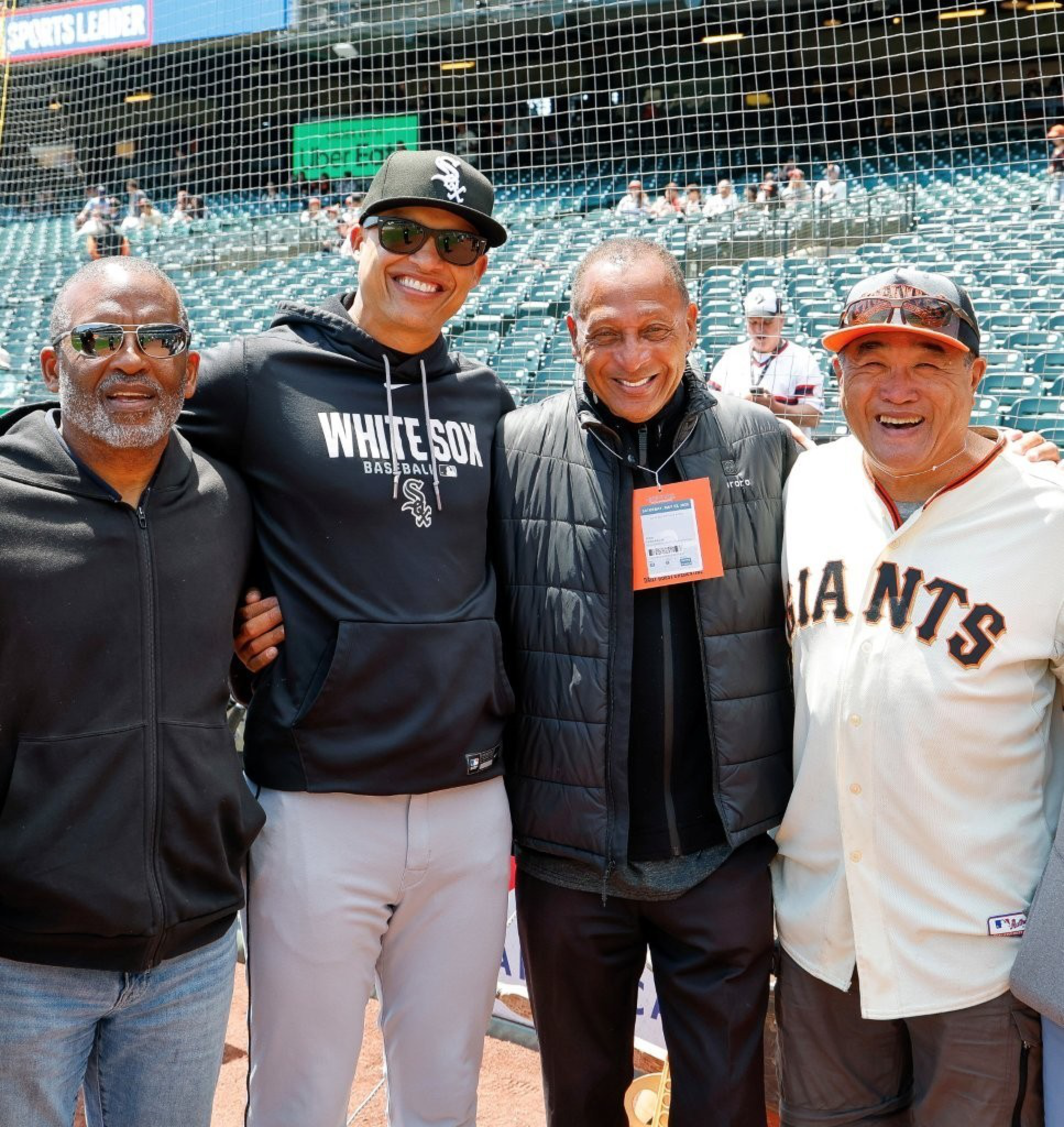 Four men stand arm in arm at a baseball stadium, two wearing baseball team attire: a White Sox hoodie and a Giants jersey.