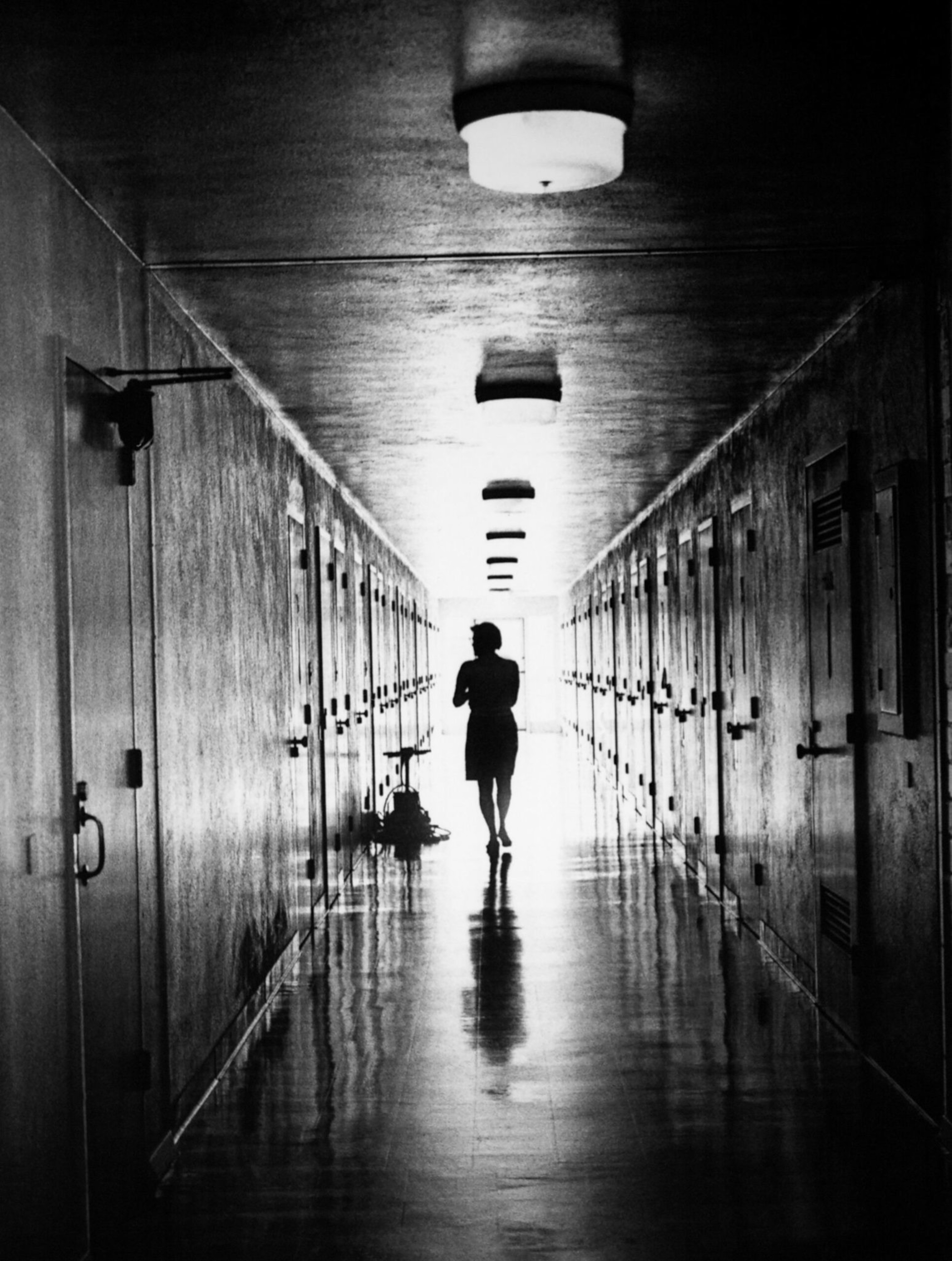 A silhouetted person walks down a long, narrow hallway lined with closed doors on both sides, illuminated by ceiling lights and bright light ahead.