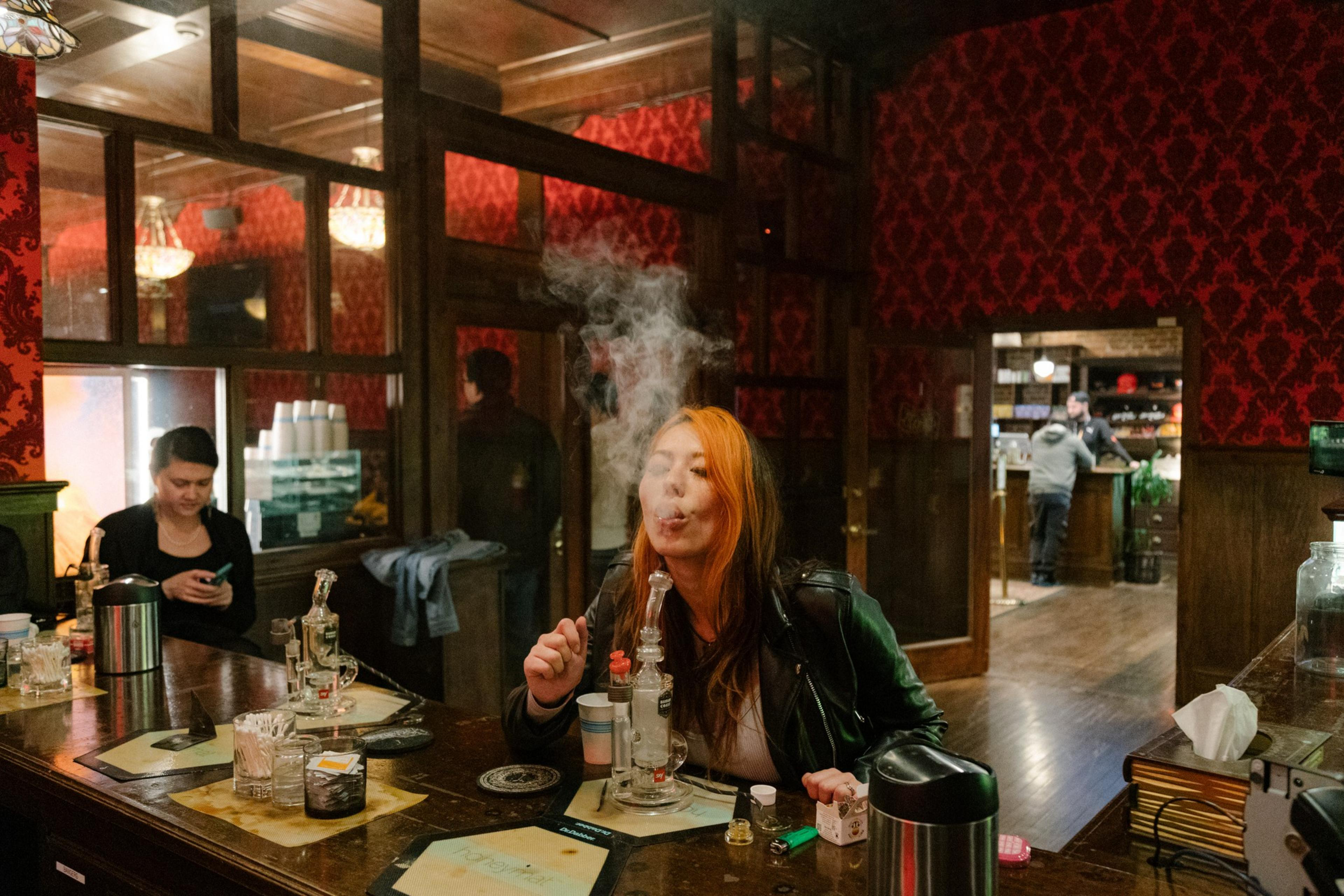 A woman blows out smoke after smoking out of a cannabis pipe in a cannabis dipsensary - some people are in the background shopping for cannabis merchandise.