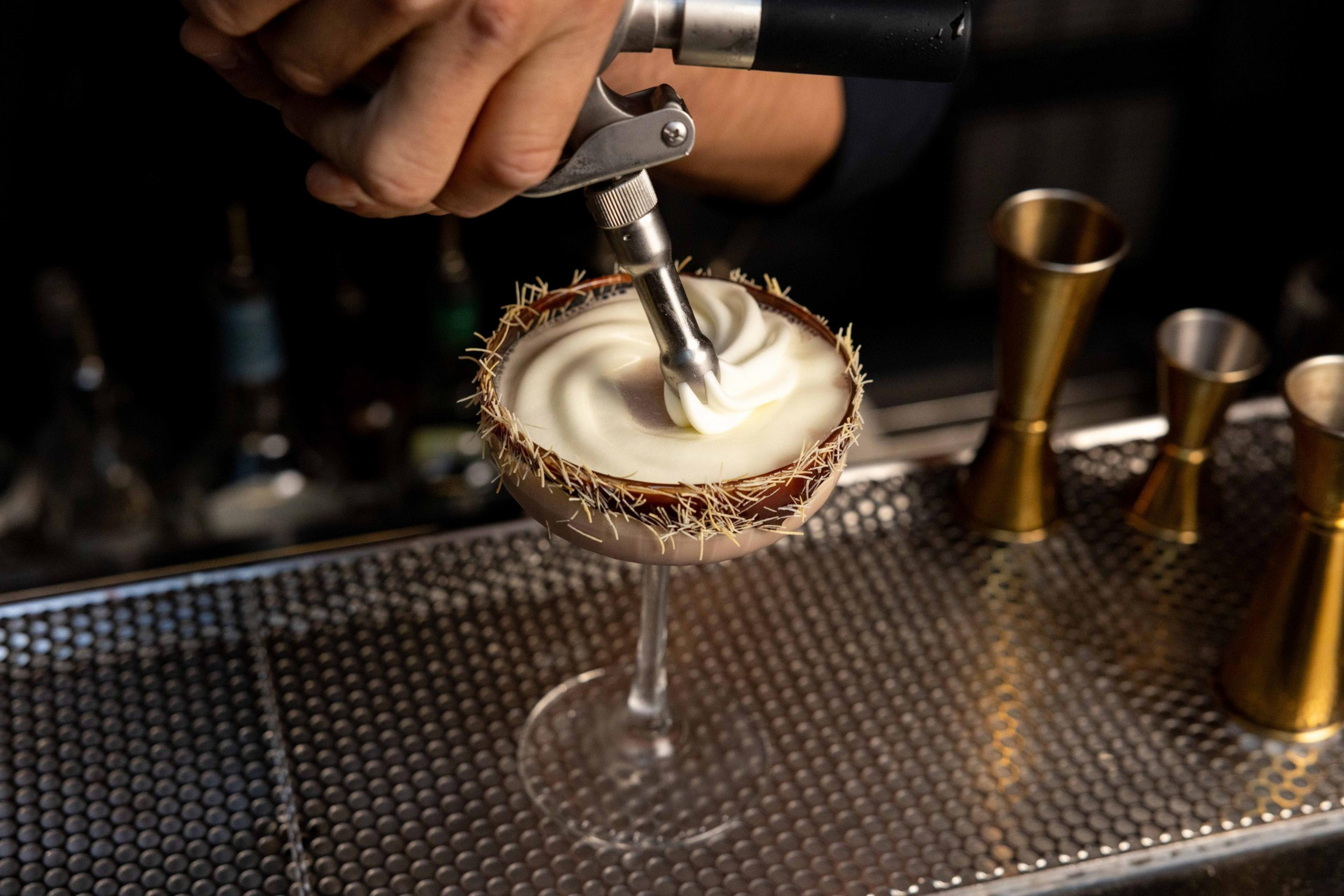 A hand is applying whipped cream onto a cocktail in a glass with a spiky rim. Several metal jiggers are on a perforated metal counter nearby.