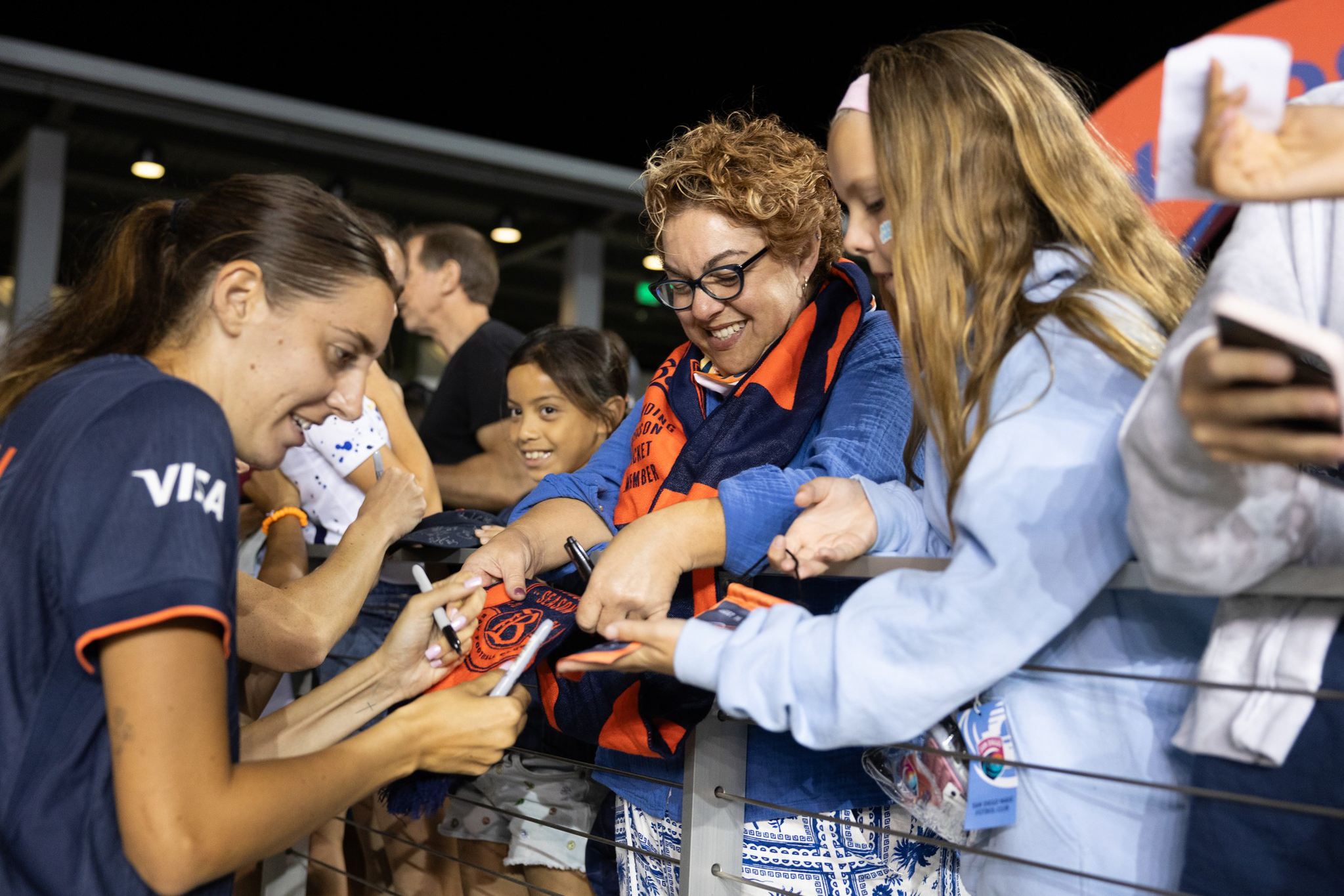 A female athlete signs autographs for excited fans, including a smiling woman in glasses and two girls, at a nighttime event behind a railing.