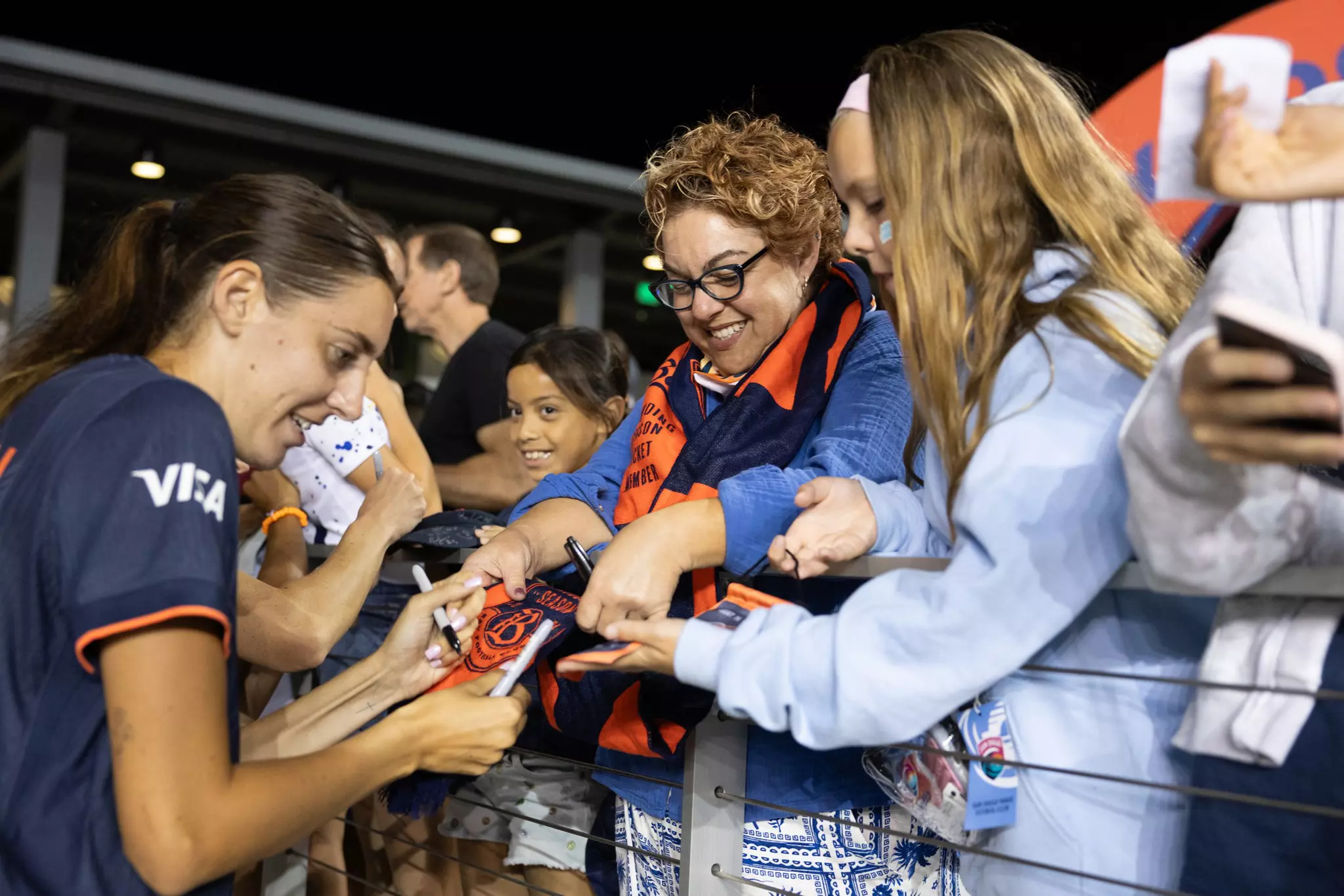 A female athlete signs autographs for excited fans, including a smiling woman in glasses and two girls, at a nighttime event behind a railing.