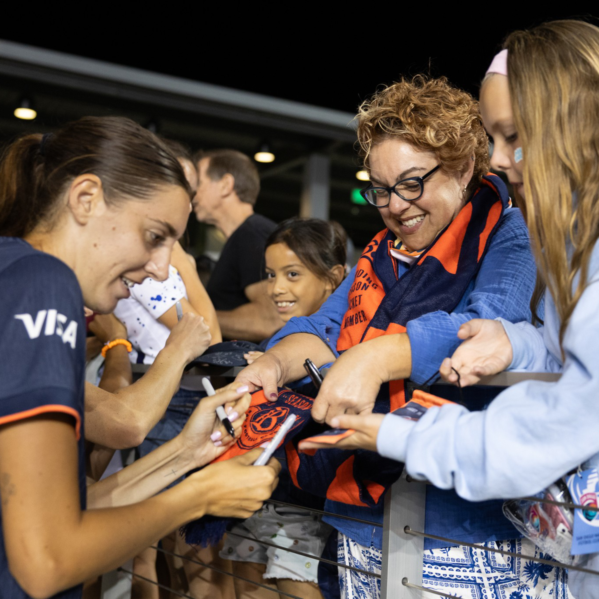 A female athlete signs autographs for excited fans, including a smiling woman in glasses and two girls, at a nighttime event behind a railing.