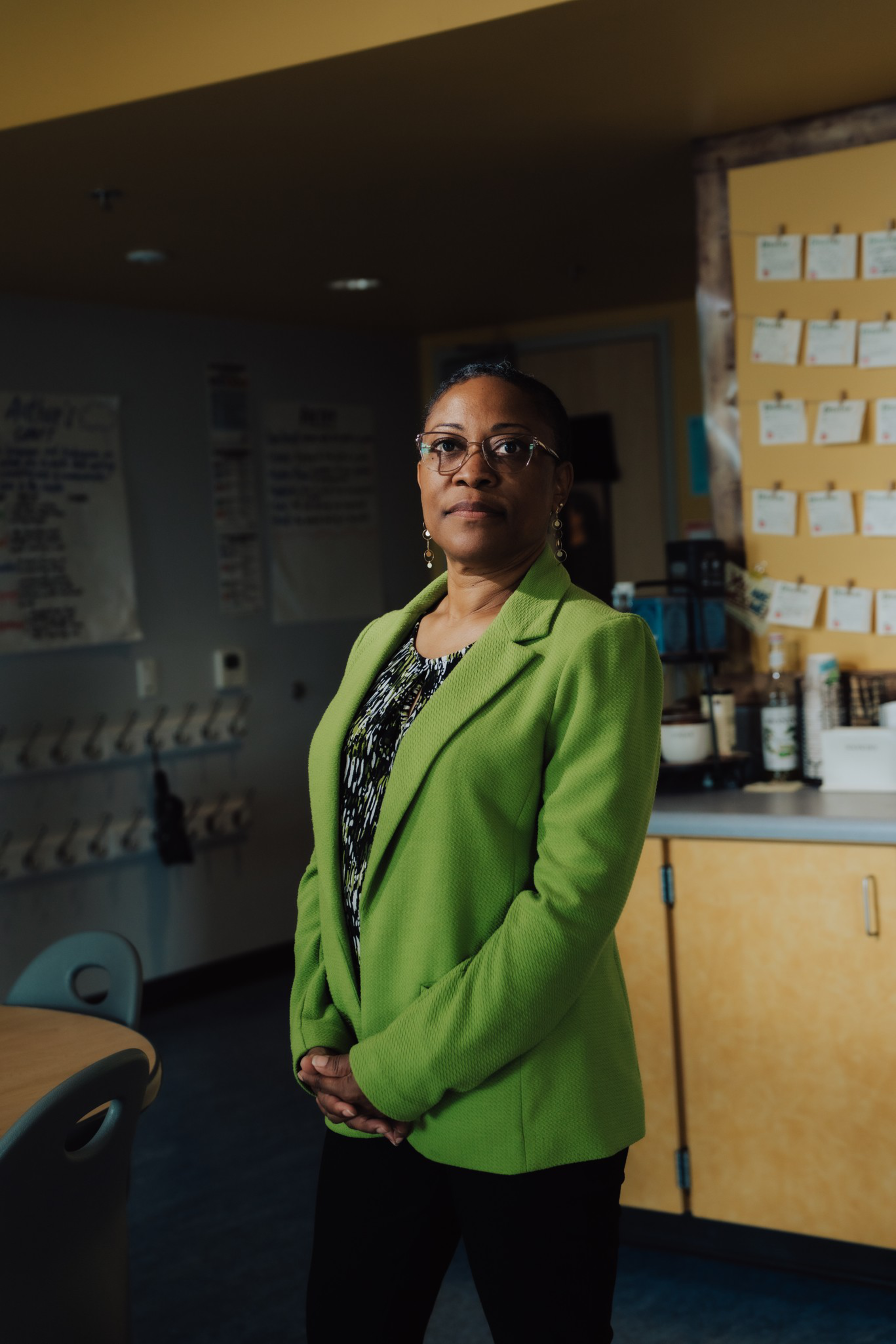 A woman wearing glasses and a green blazer stands with hands clasped in a room with chairs, a table, cabinets, and notes pinned on a bulletin board.