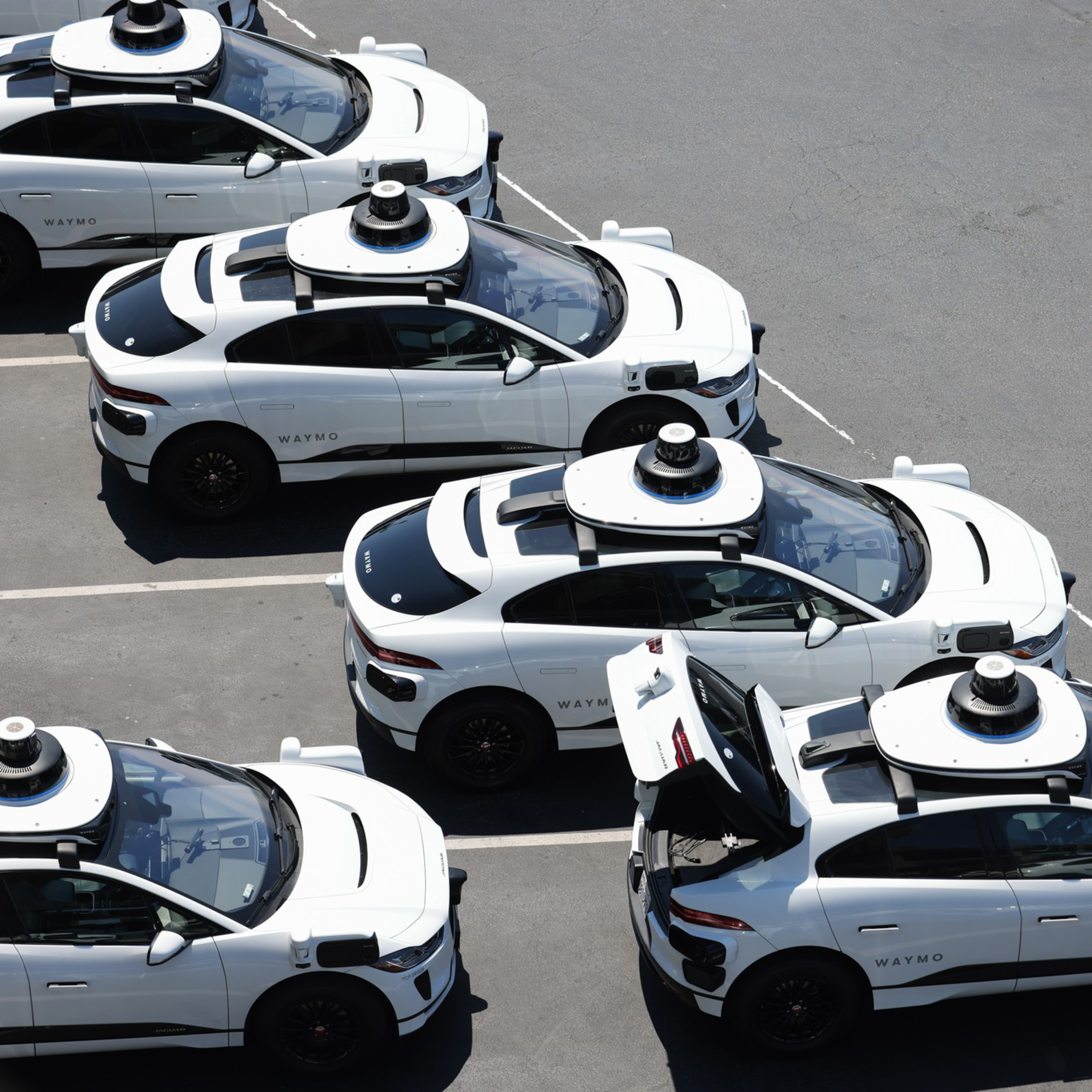 The image shows several white Waymo self-driving cars parked in a lot. They have dome-like sensors on their roofs, and one car's trunk is open.