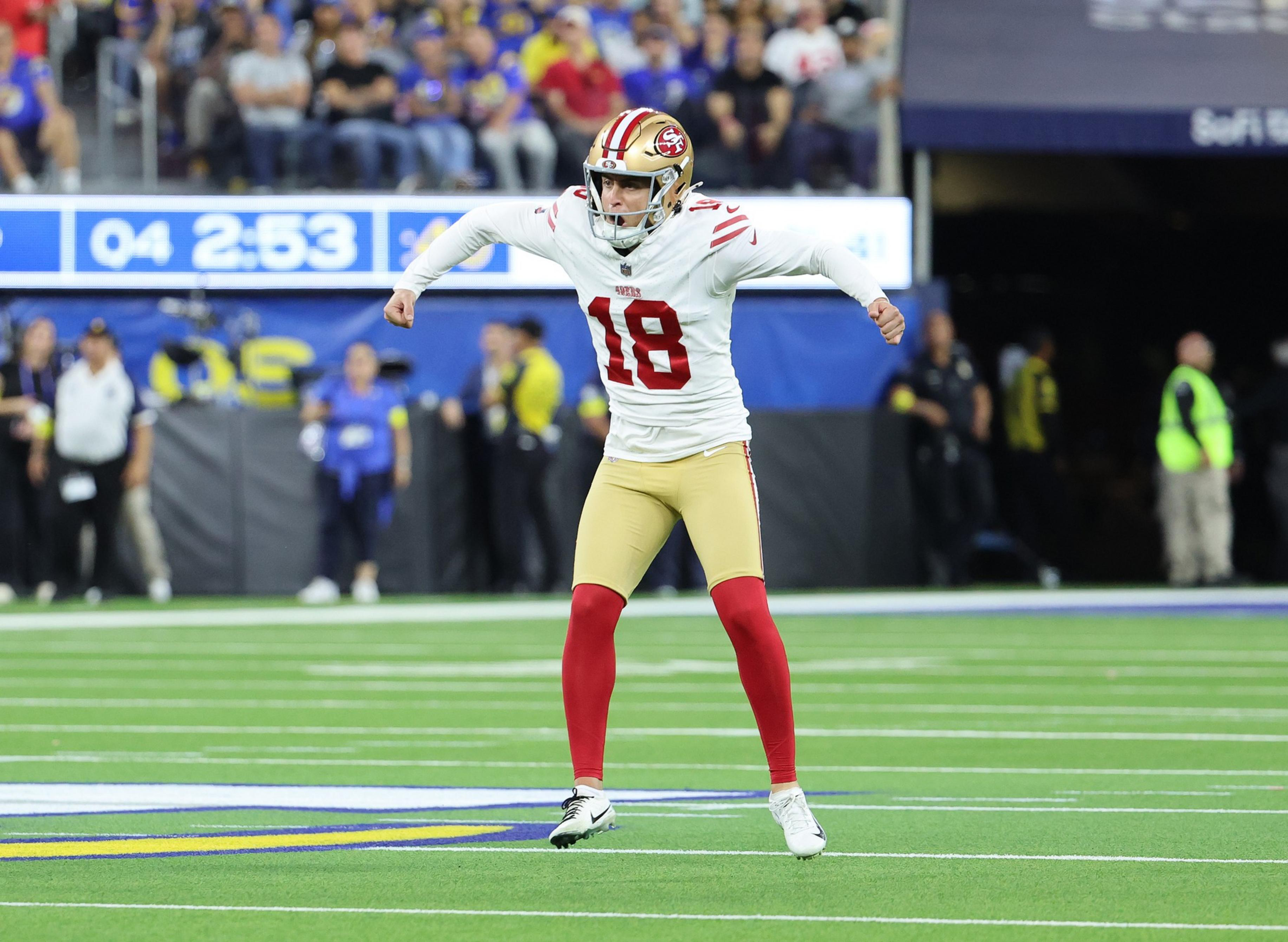 A football player in a white and red uniform with the number 18 is jumping or celebrating on a green field with an audience in the background.
