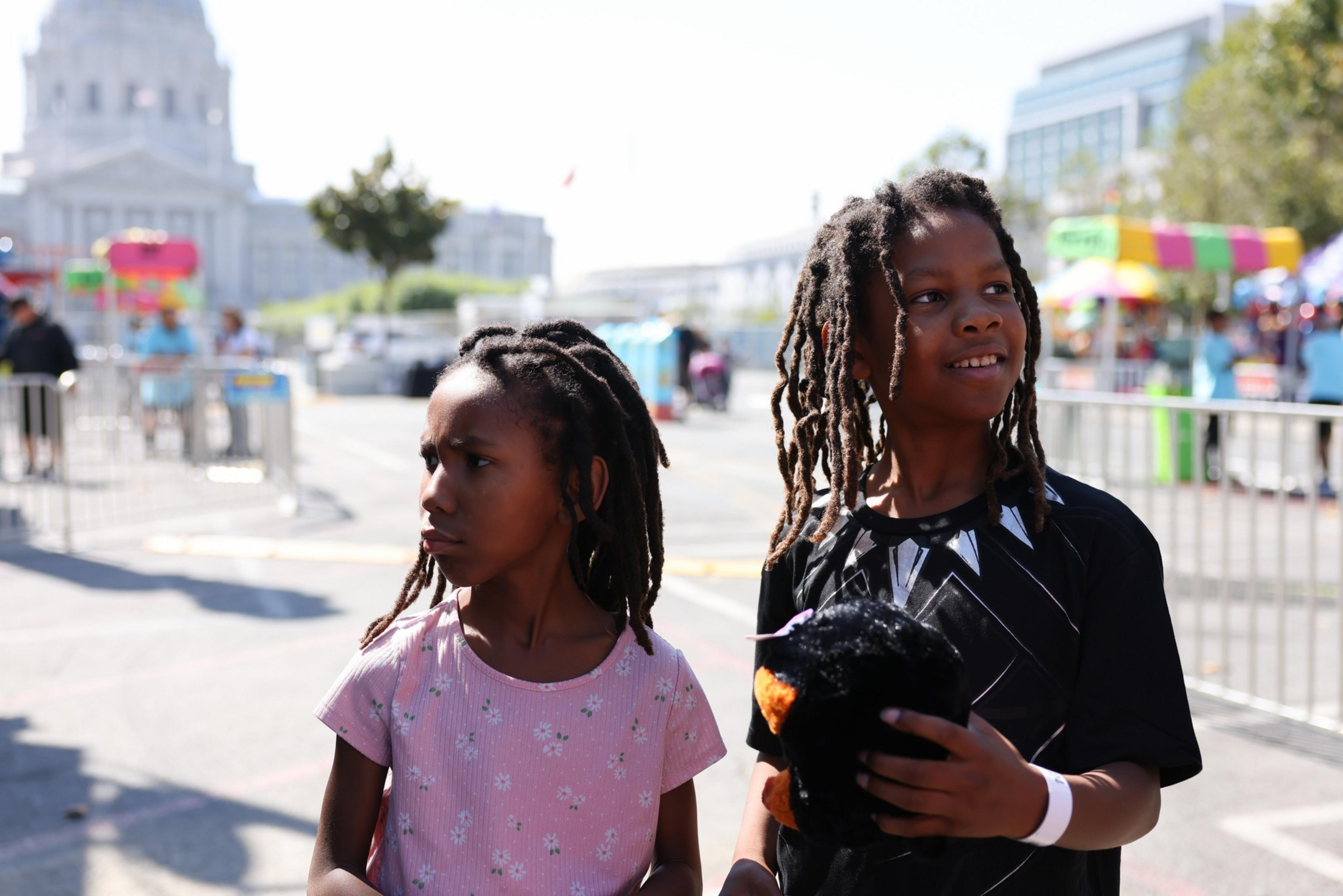 Two children with braided hair stand outdoors, one in a pink shirt looking to the side, the other in a black shirt smiling and holding a black stuffed animal.