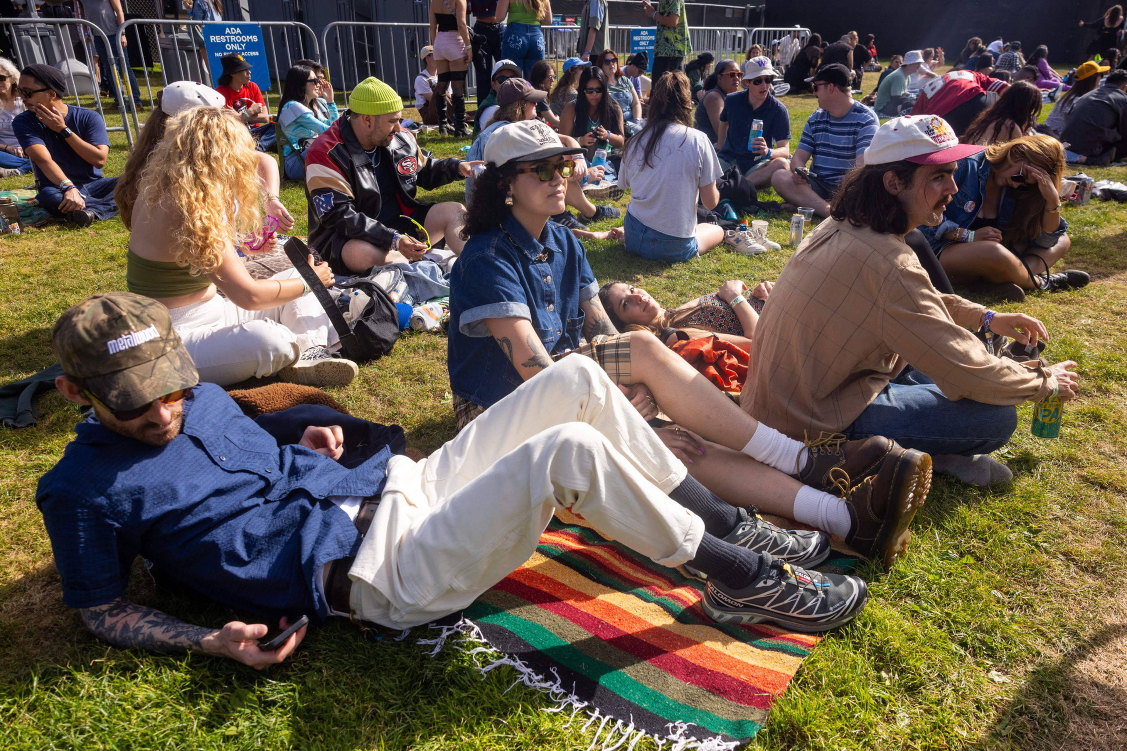 People sit and lie on the grass at an outdoor event, some on blankets, enjoying the sunny weather and socializing in a relaxed atmosphere.