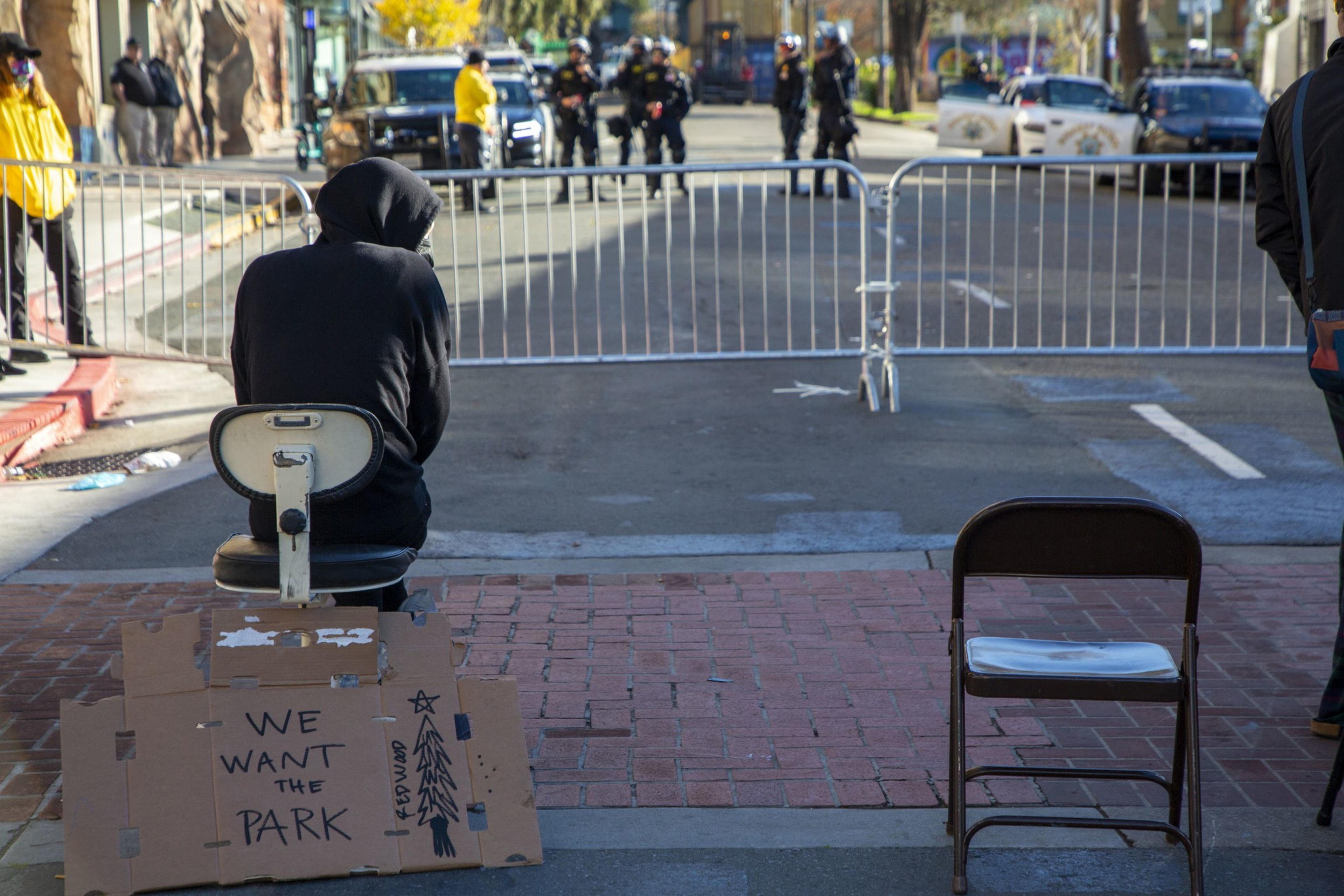 A person wearing a dark hooded sweatshirt sits in a chair in front of police on the other side of a metal barricade; behind the person, a cardboard sign reads &quot;we want the park.&quot;