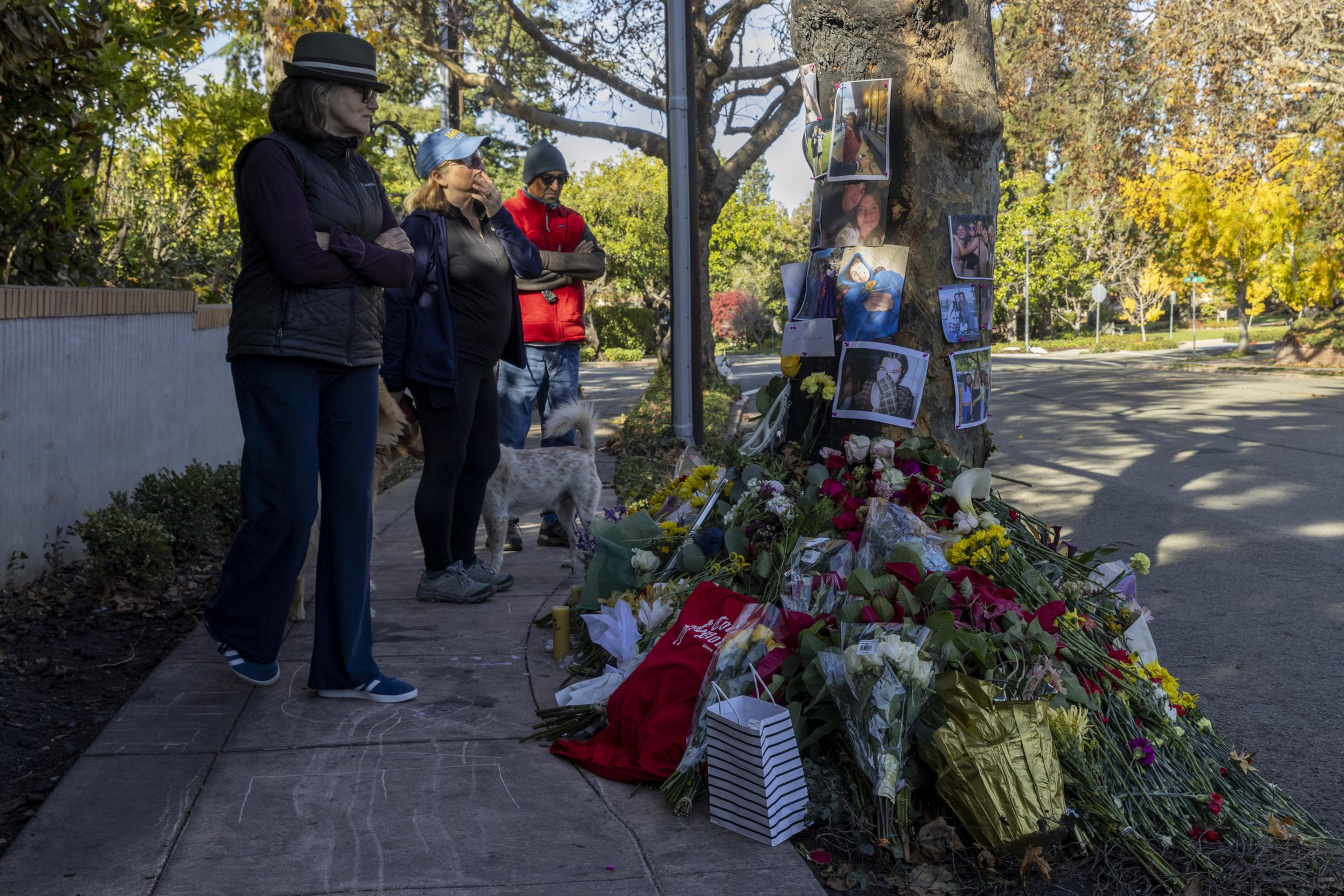 A small group of people stands solemnly on a sidewalk, observing a memorial with flowers and photos arranged against a tree. A dog is present nearby.