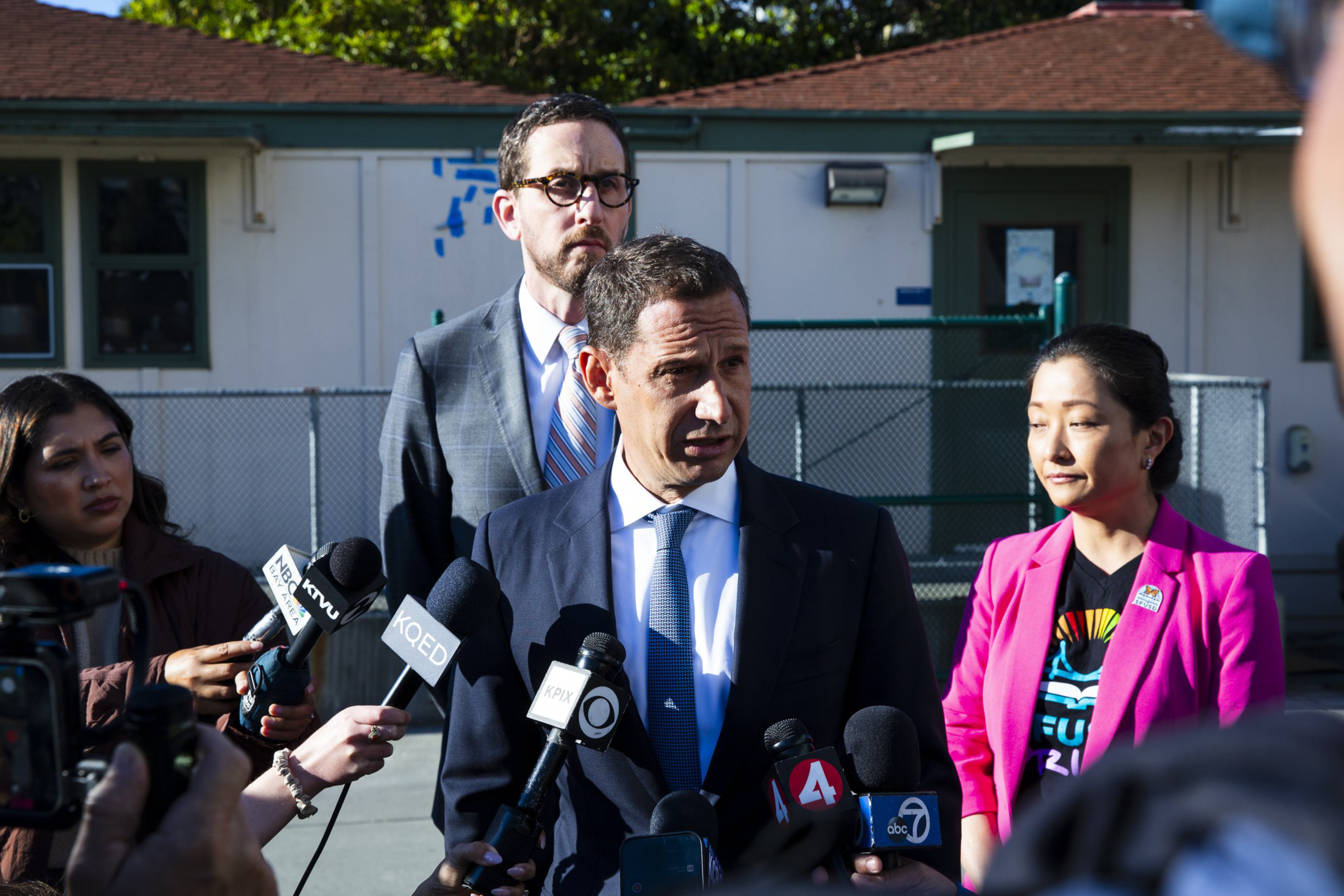 A man in a suit and blue tie speaks into multiple microphones outdoors, flanked by two people, one in a pink blazer, with a building and fence behind them.