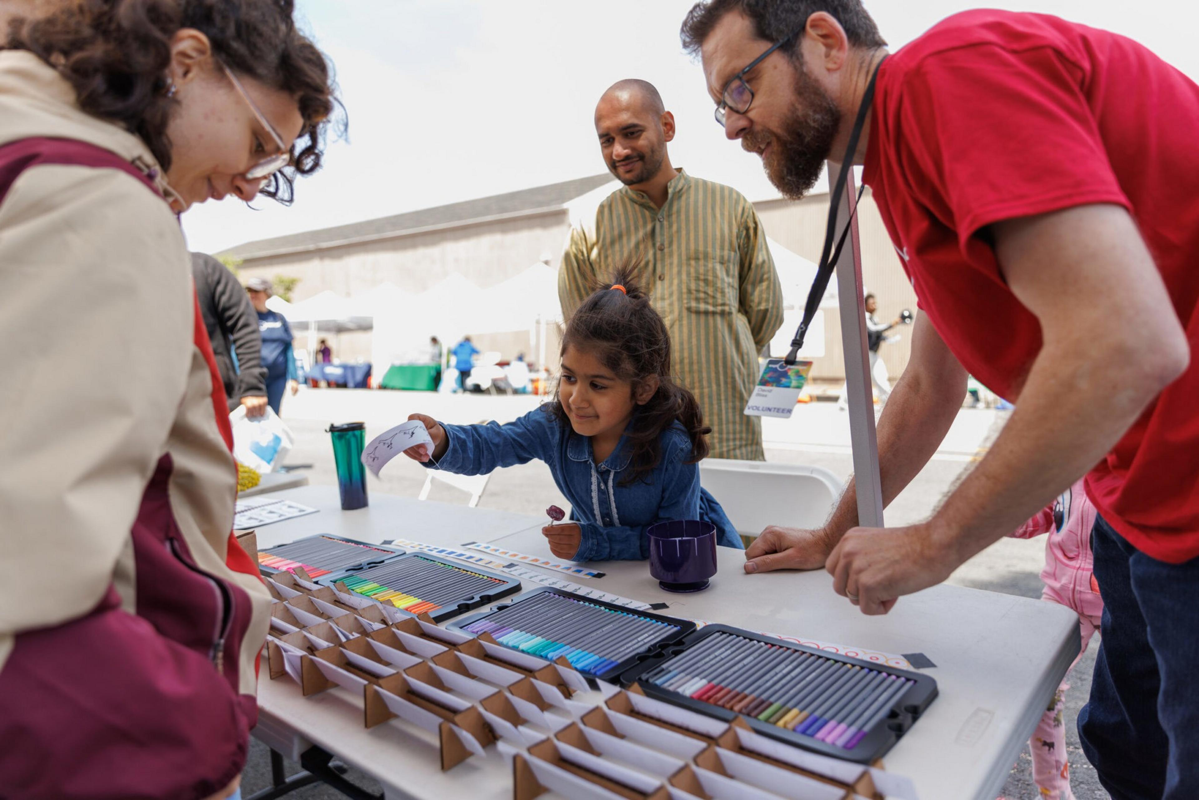 A child hands a drawing to a man at a colorful outdoor art booth as adults watch.