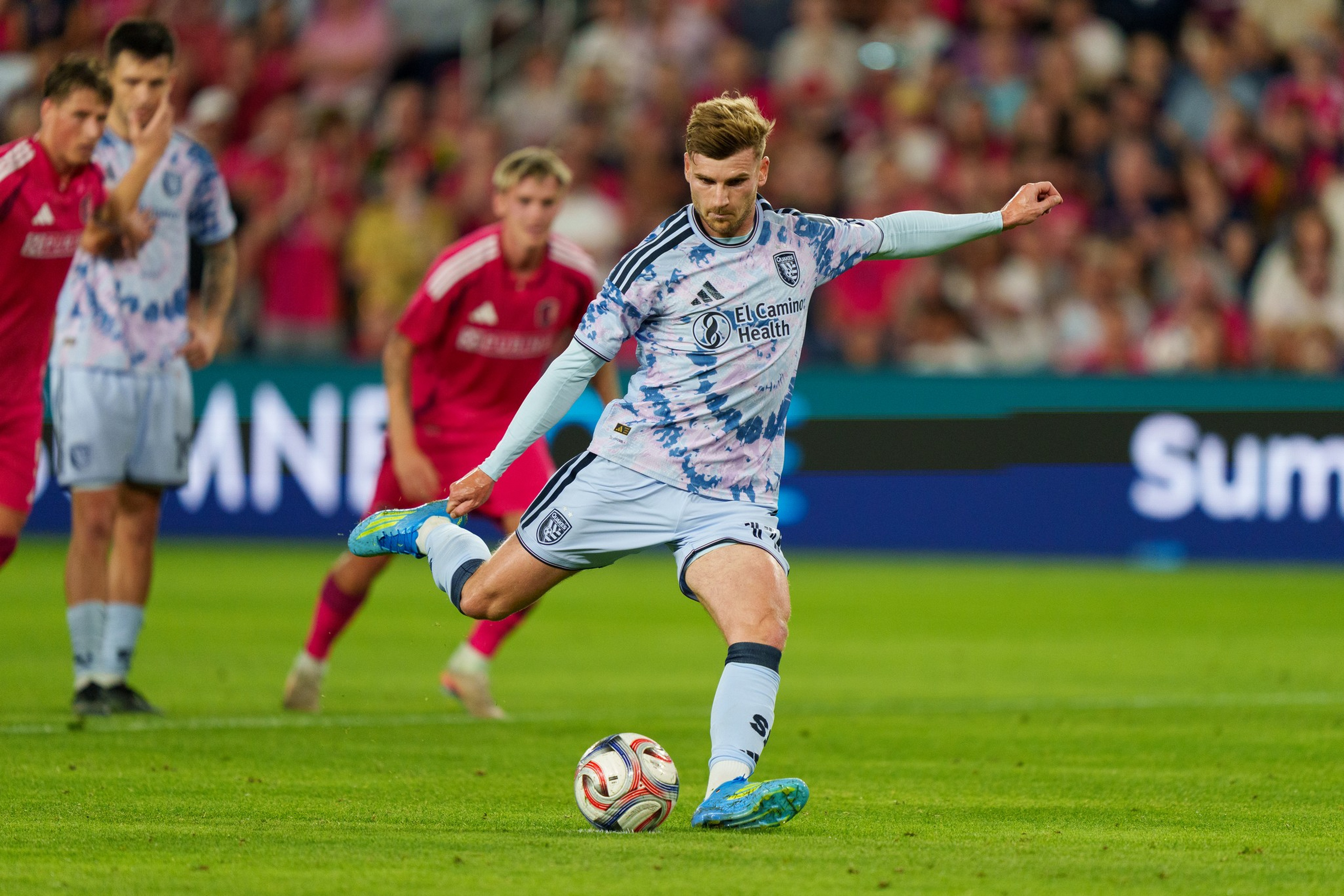 A soccer player in a light blue tie-dye uniform is about to kick a ball, while players in red uniforms watch in the background on a green field.