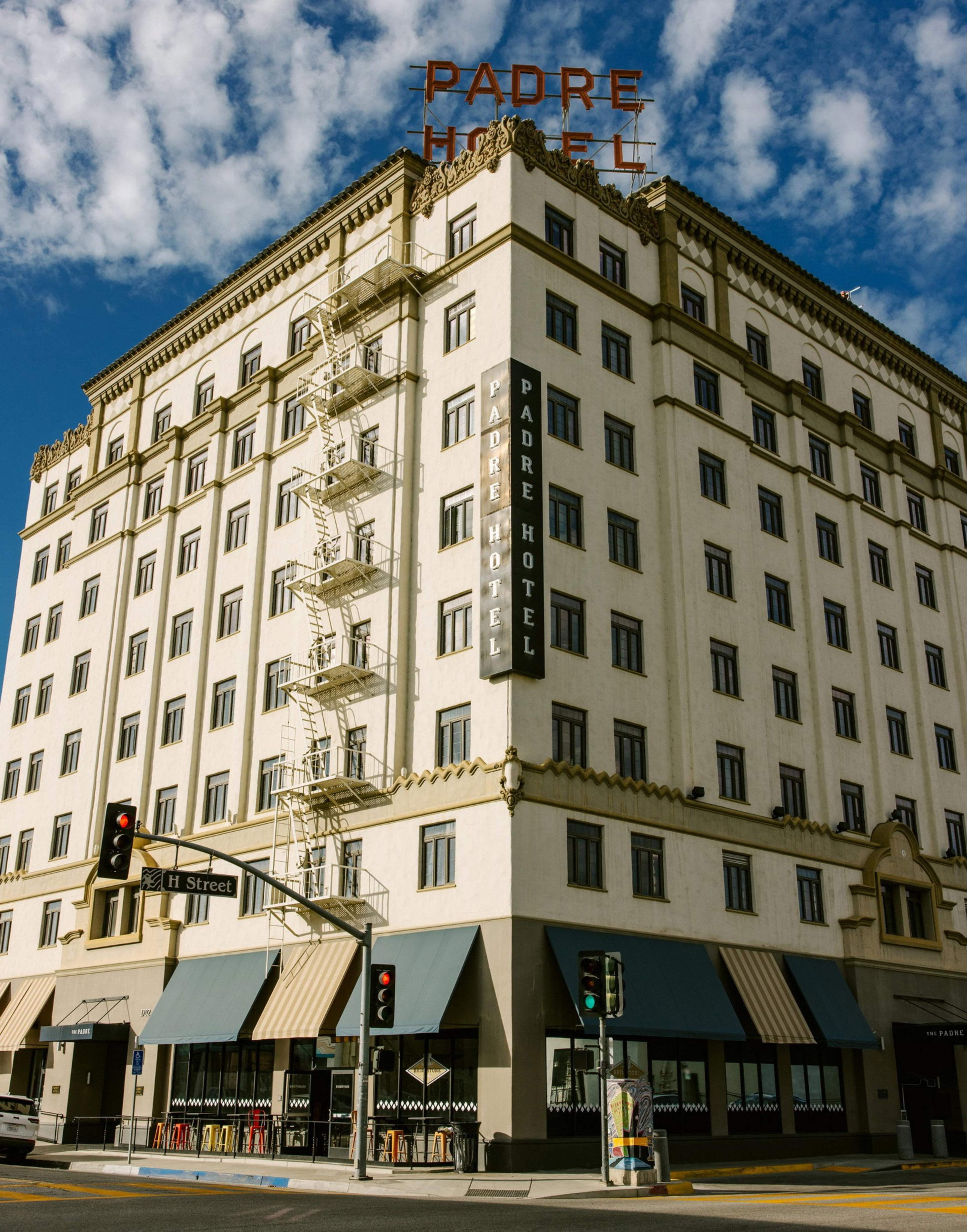 A white, multi-story building labeled &quot;Padre Hotel&quot; features a vintage design with a rooftop sign, striped awnings, and fire escape stairs on a sunny day.