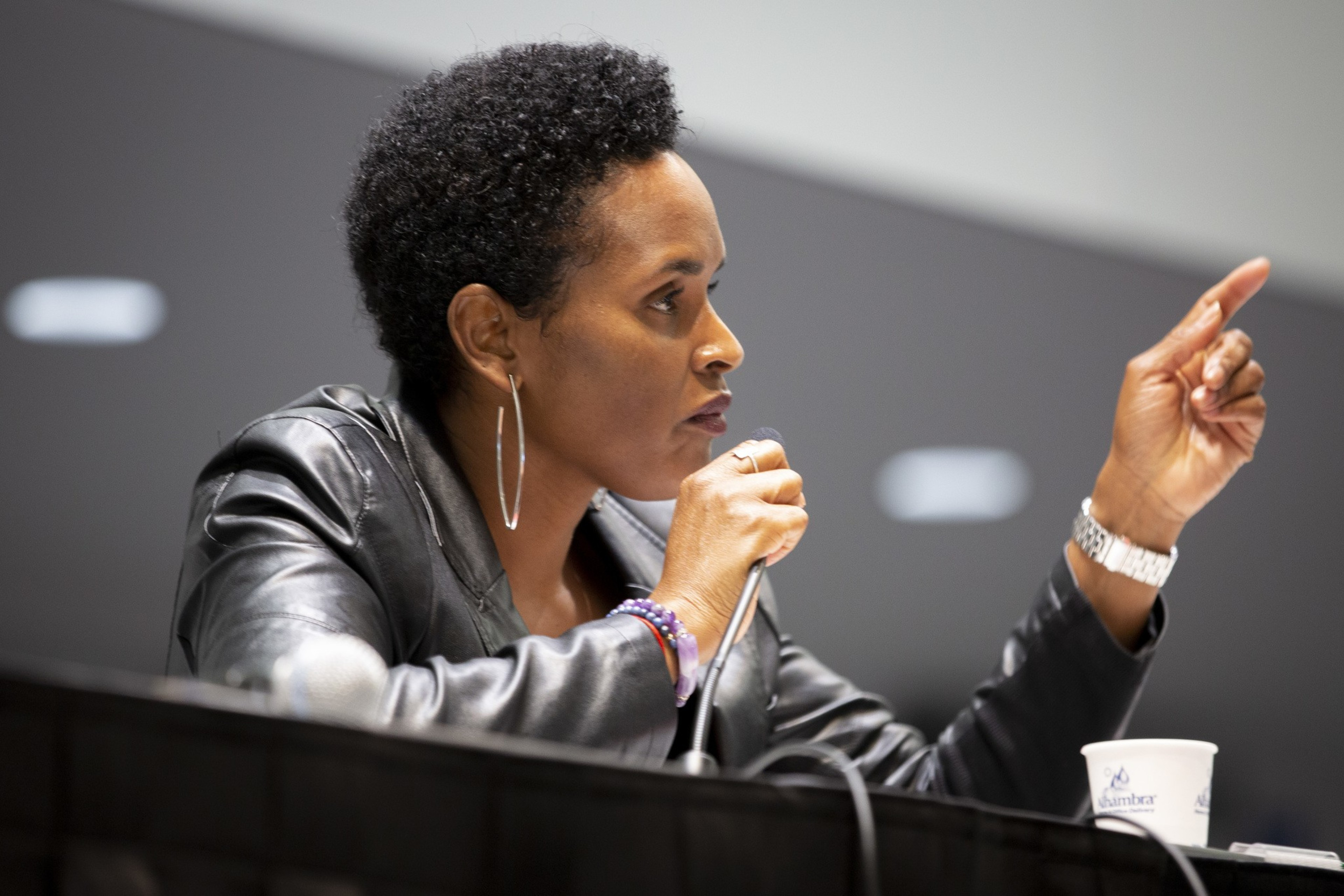 A woman with short hair is speaking into a microphone while pointing with her finger. She wears a leather jacket, hoop earrings, and a watch, sitting at a table.