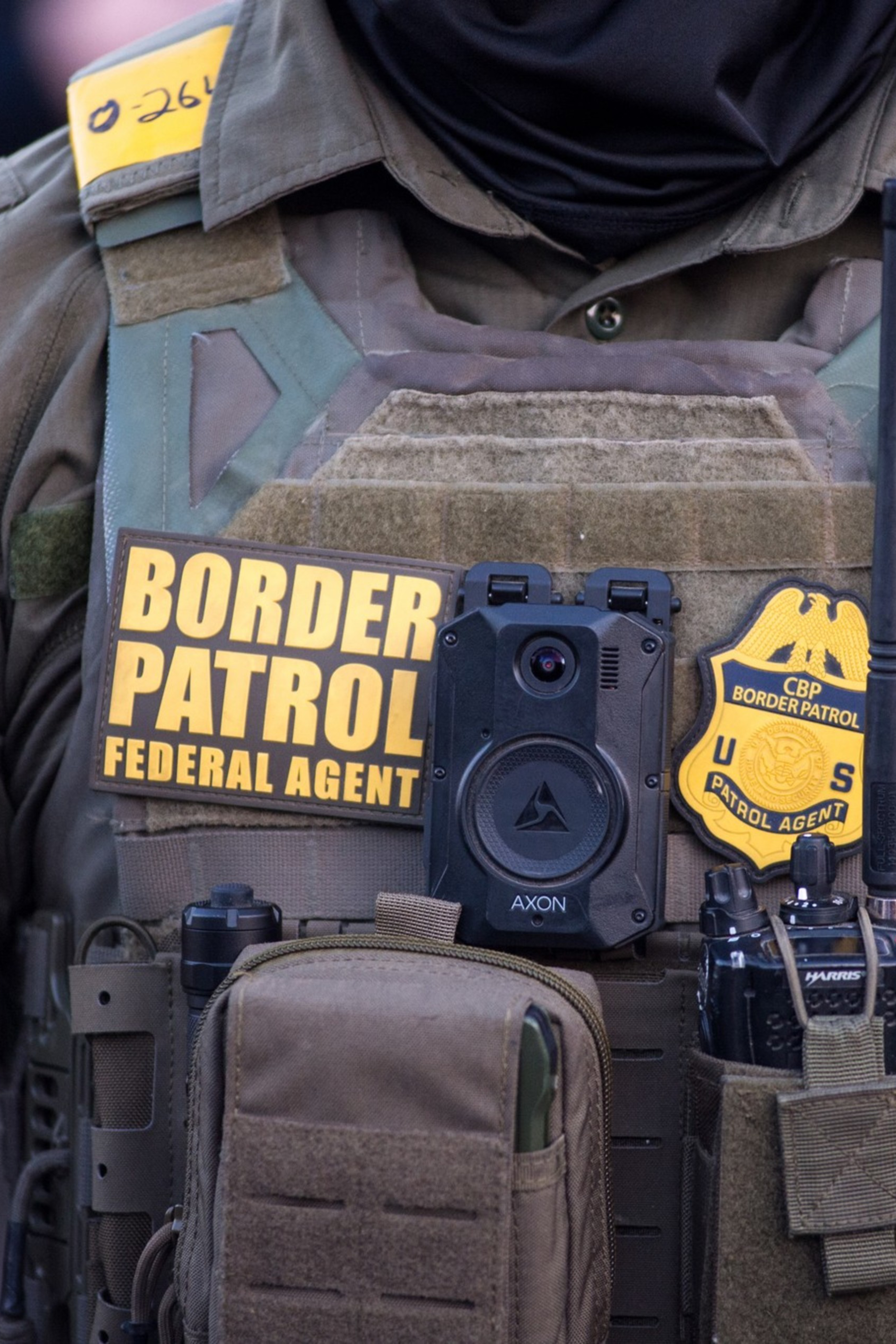 A Border Patrol agent wears tactical gear including a body camera, radio, badges, and patches identifying them as a federal agent.