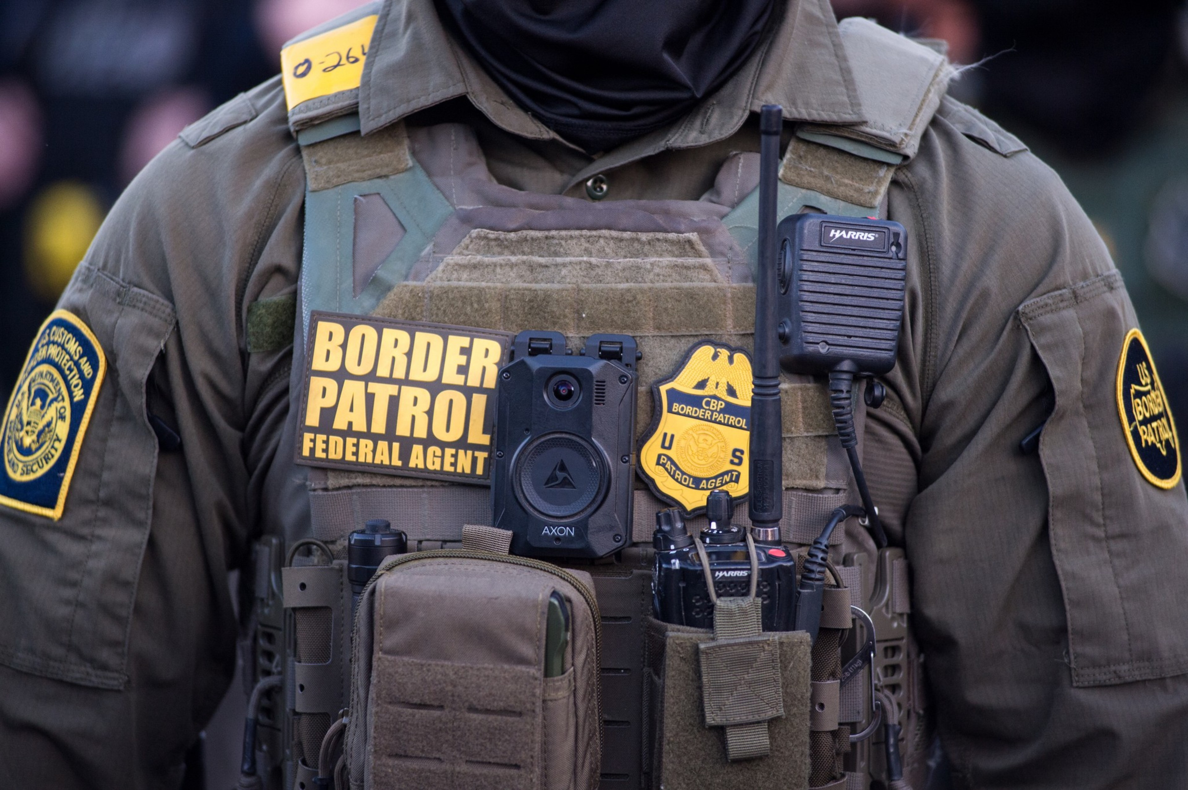 A Border Patrol agent wears tactical gear including a body camera, radio, badges, and patches identifying them as a federal agent.