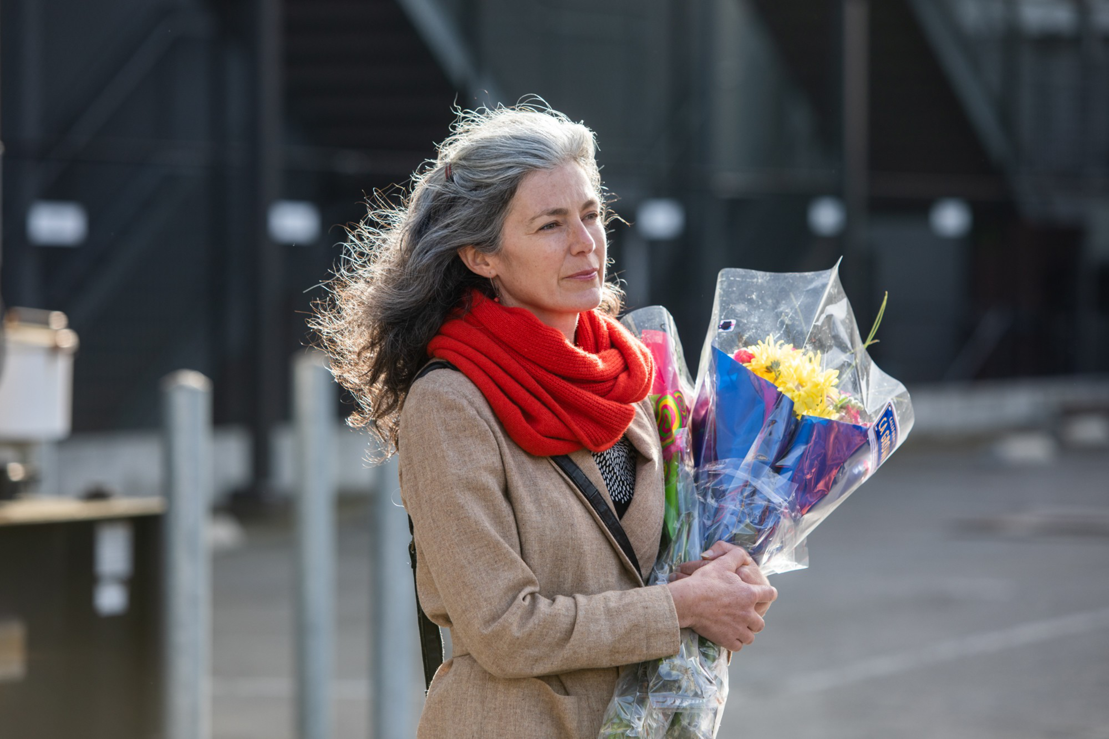 A gray-haired woman in a beige coat and red scarf holds two bouquets of flowers wrapped in clear plastic, standing outdoors on a gray day.