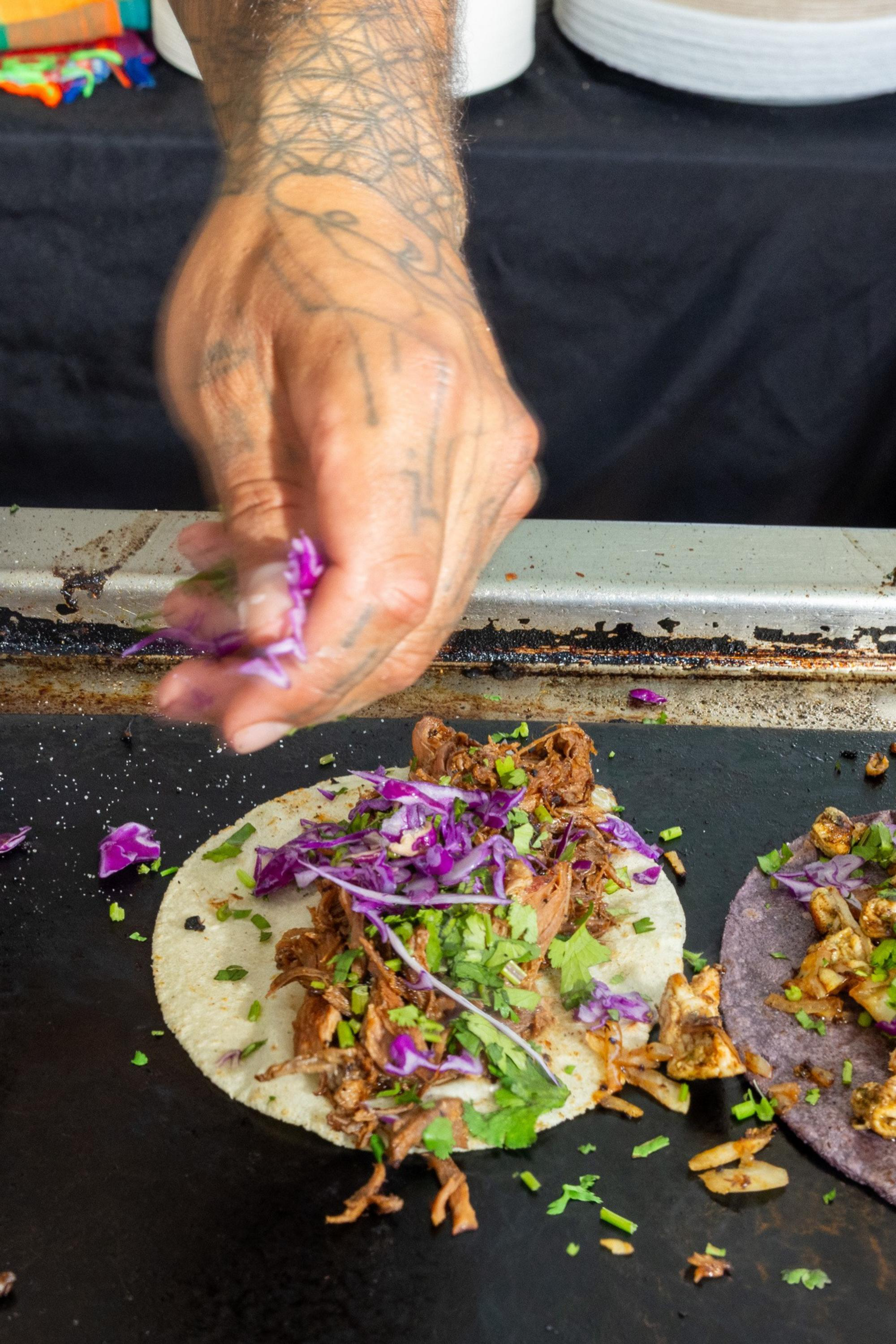 A tattooed hand is sprinkling chopped purple cabbage and green herbs onto a taco filled with shredded meat on a flat cooking surface.