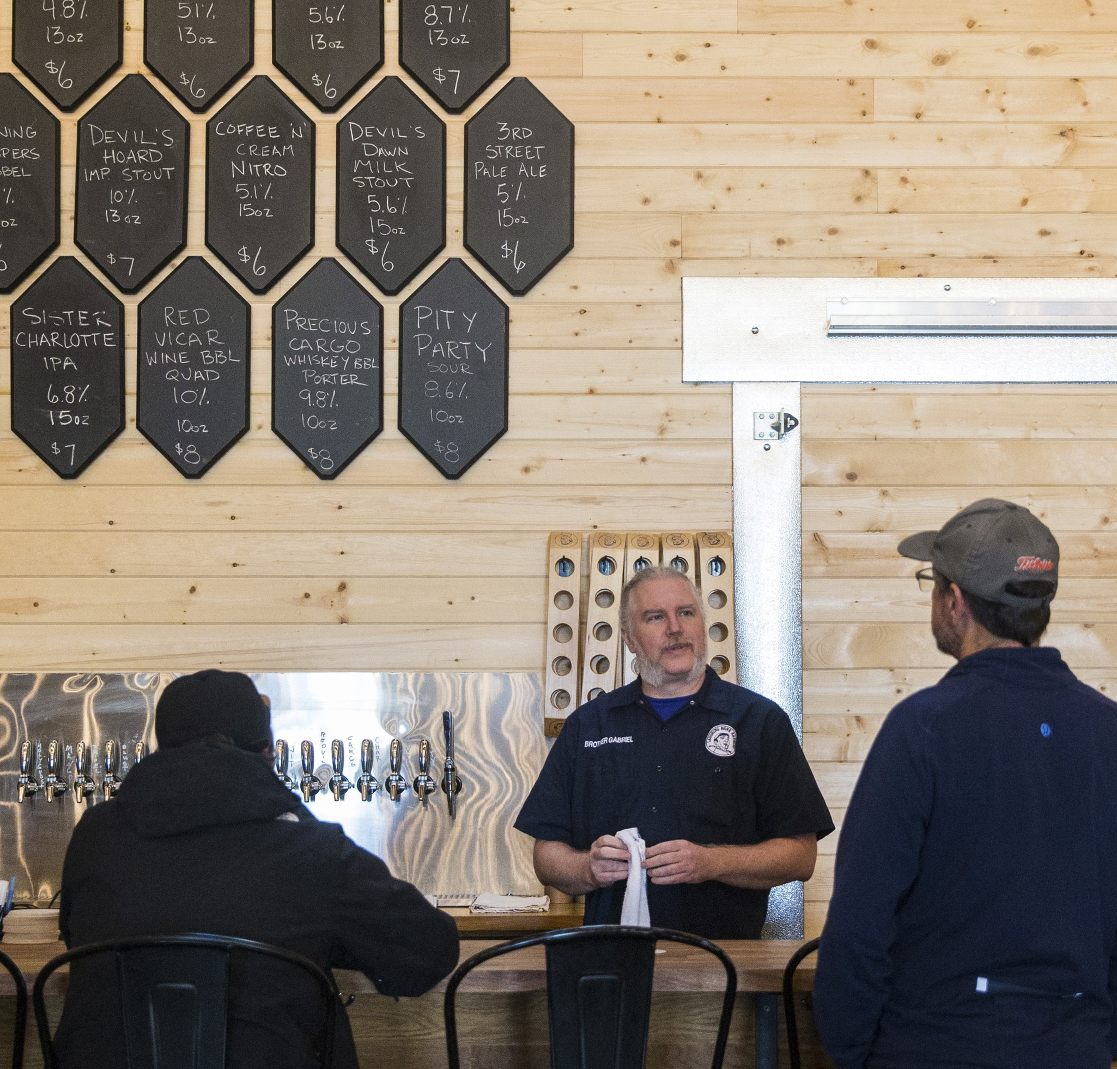 A bartender stands behind a wooden bar with taps, talking to two customers, with a blackboard menu listing beers and prices on the wall.