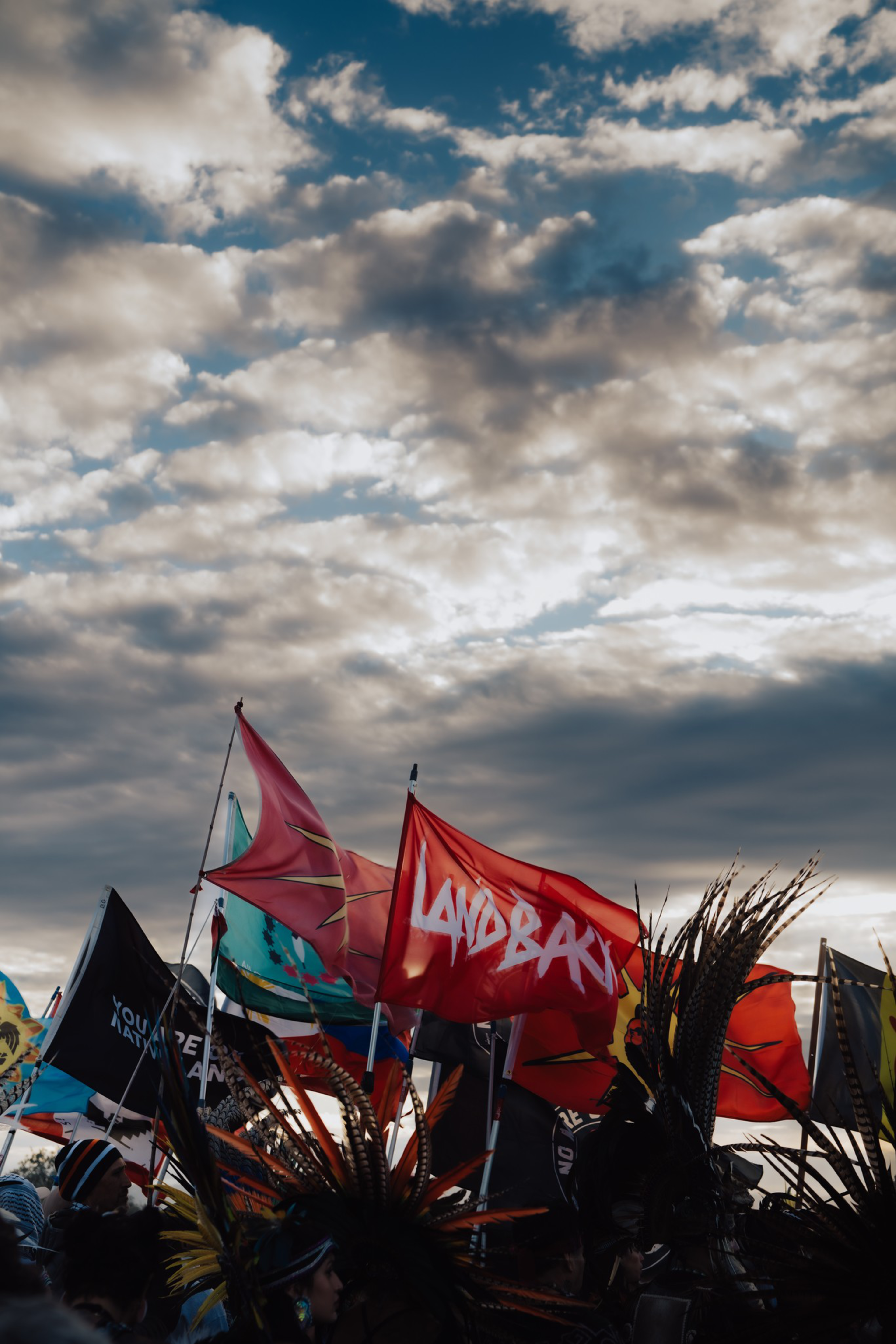 Colorful flags and people wearing feathered headdresses gather under a dramatic, cloudy sky during a vibrant outdoor event.