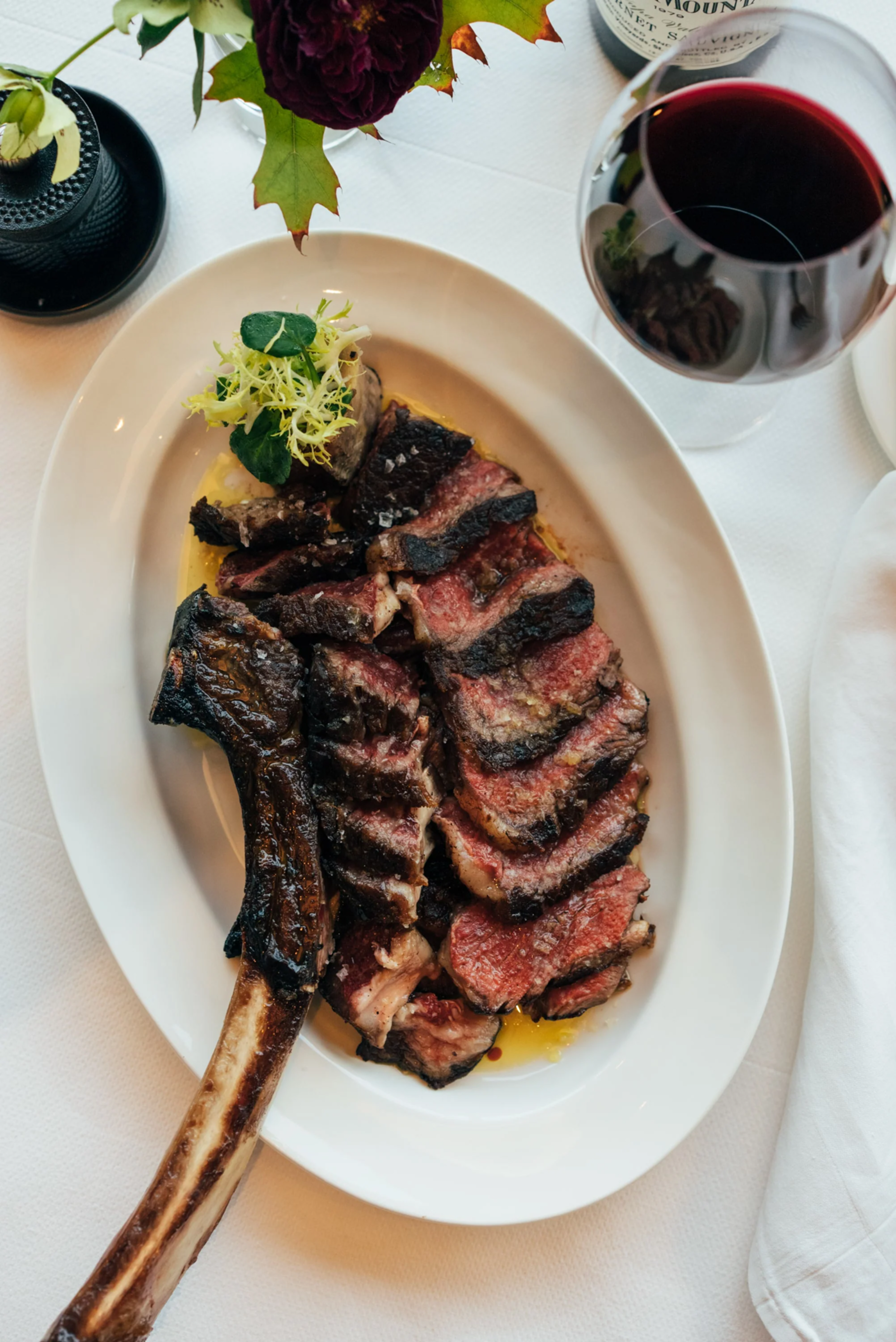 A sliced, juicy tomahawk steak is presented on a white plate, garnished with greens. A glass of red wine and a dark flower vase are nearby on a white tablecloth.