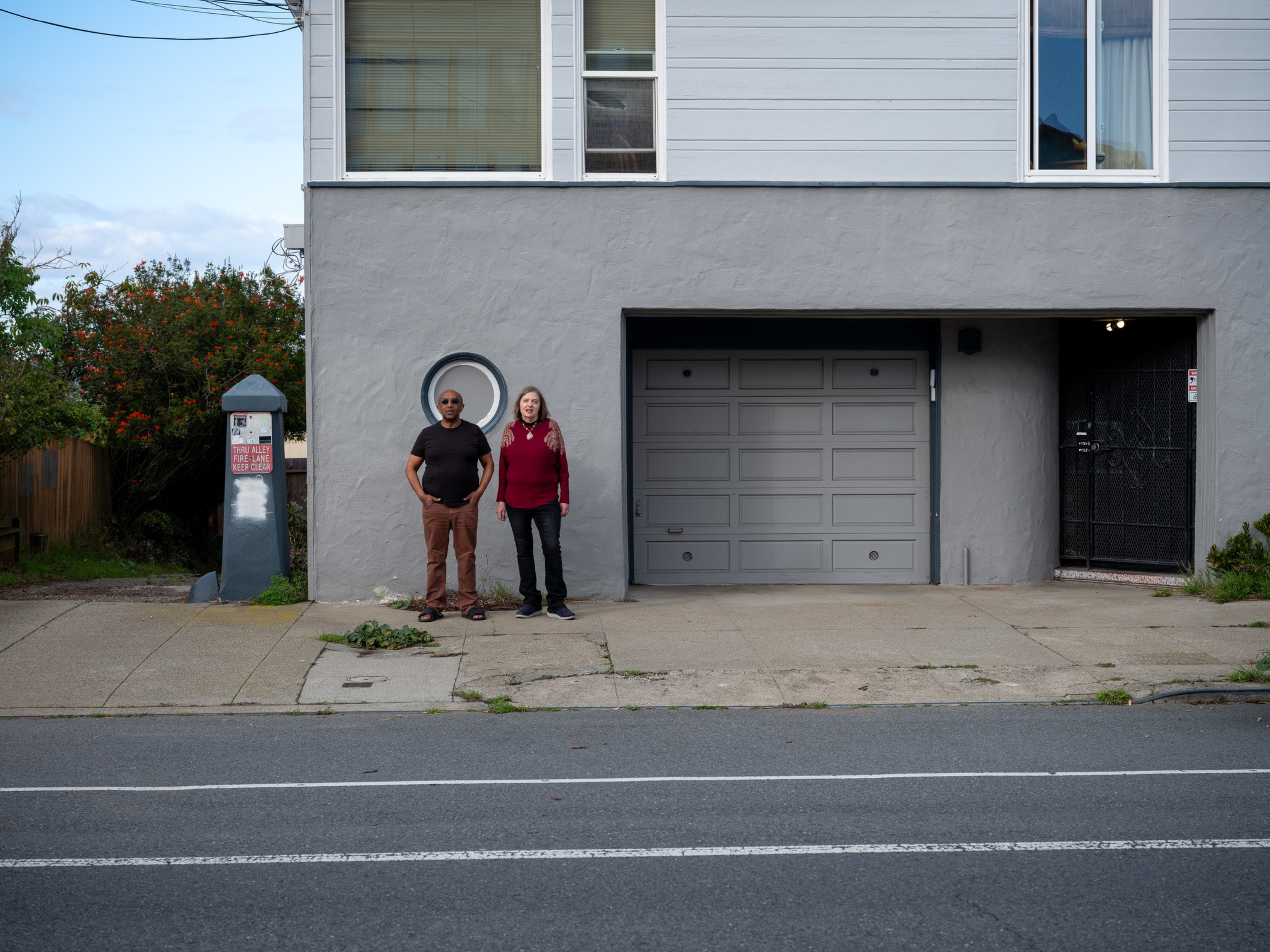 A man and woman stand on a sidewalk in front of a gray building with a garage door, circular window, and a black gated entrance.