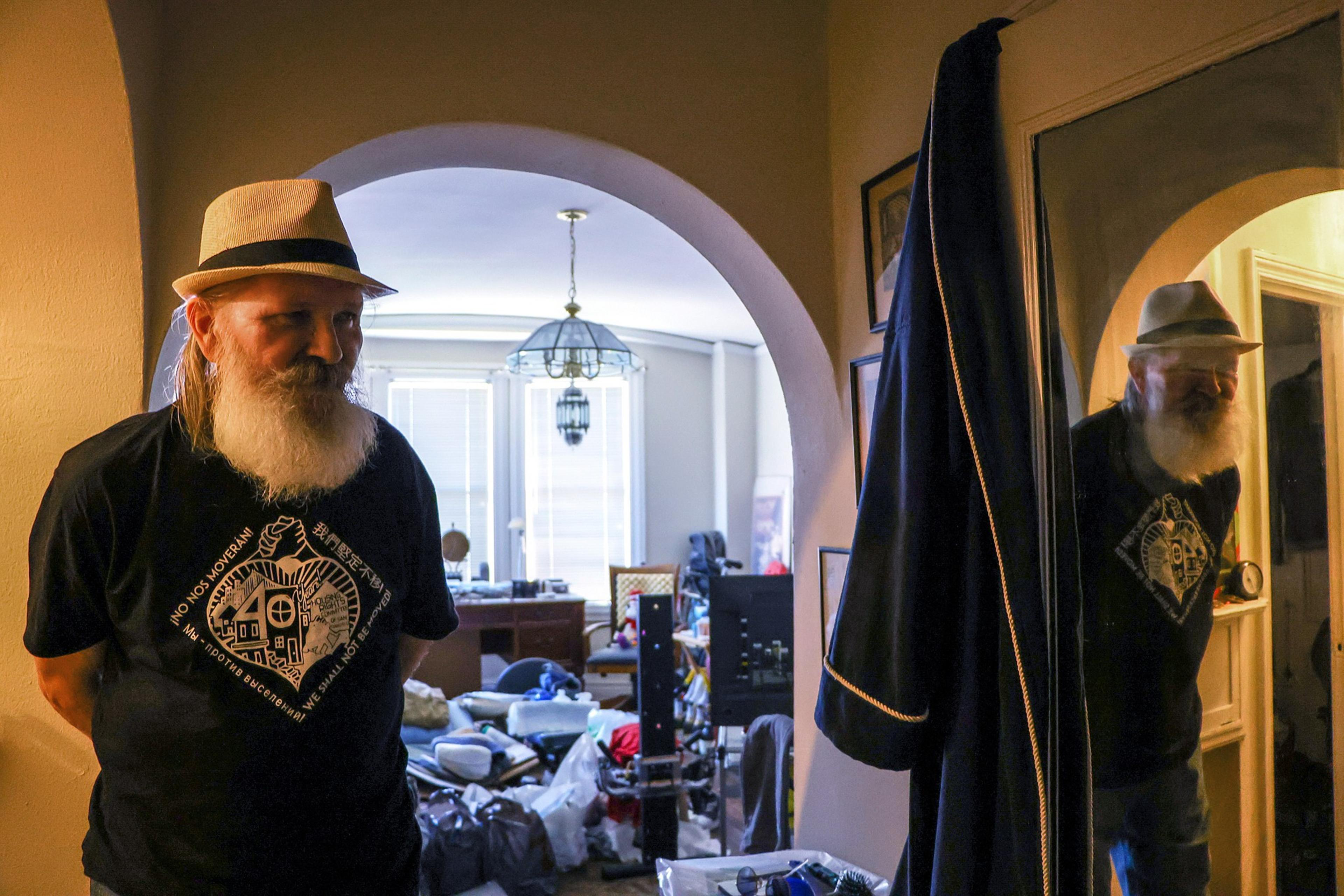 An elderly man with a white beard and hat stands in a dimly lit room with a cluttered, sunlit living area visible through an archway behind him.